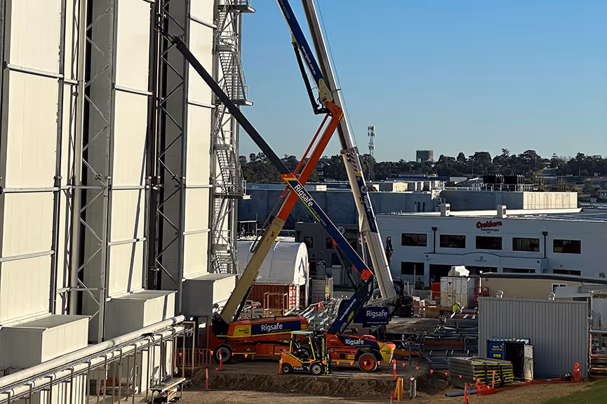 Construction site with multiple Rigsafe-branded cranes and a truck with elevated booms near a large building under clear blue sky.