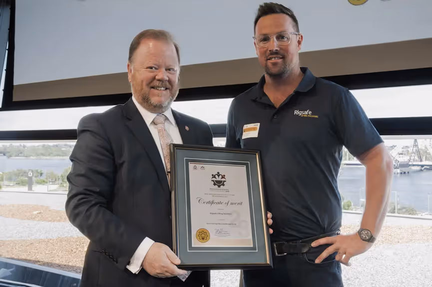 Two men standing indoors, one in a suit and the other in a Rigsafe polo shirt, holding a framed certificate of merit.