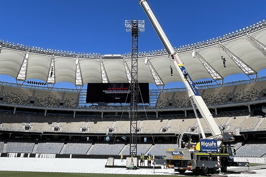 Crane and workers installing or maintaining a tall black metal structure in a large stadium with empty seats under a clear blue sky.