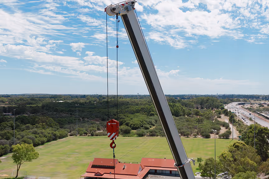 Tall crane lifting a red hook over a building with green fields, trees, and a distant highway under a partly cloudy sky.