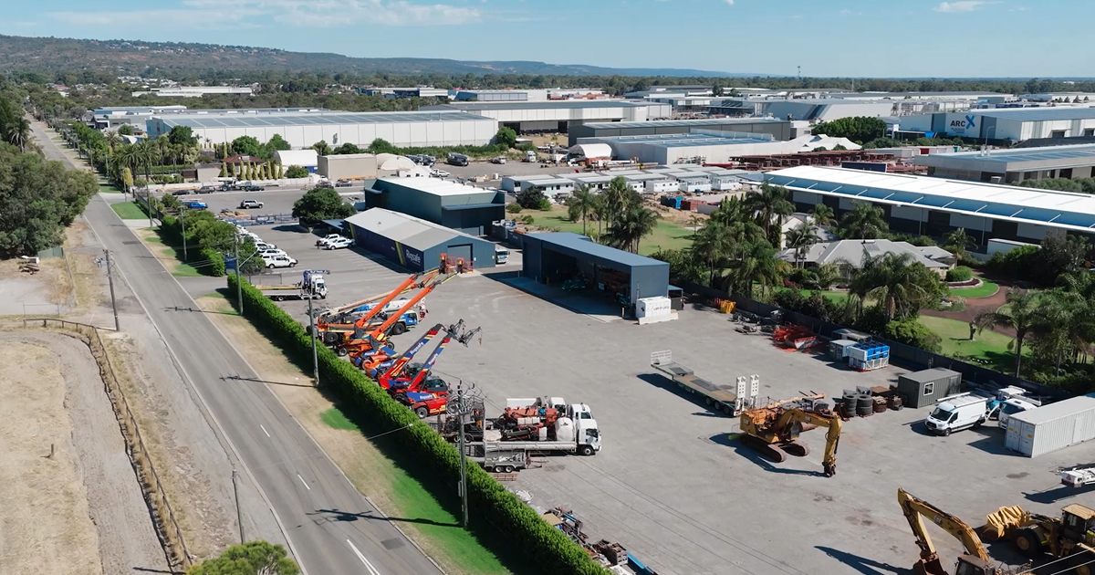 Aerial view of an industrial yard with heavy machinery, trucks, orange cranes, and multiple warehouses surrounded by greenery and palm trees.