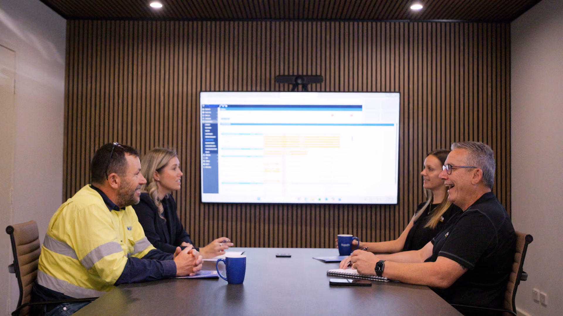 Four people sitting at a conference table in a meeting room with a screen showing data on the wall.