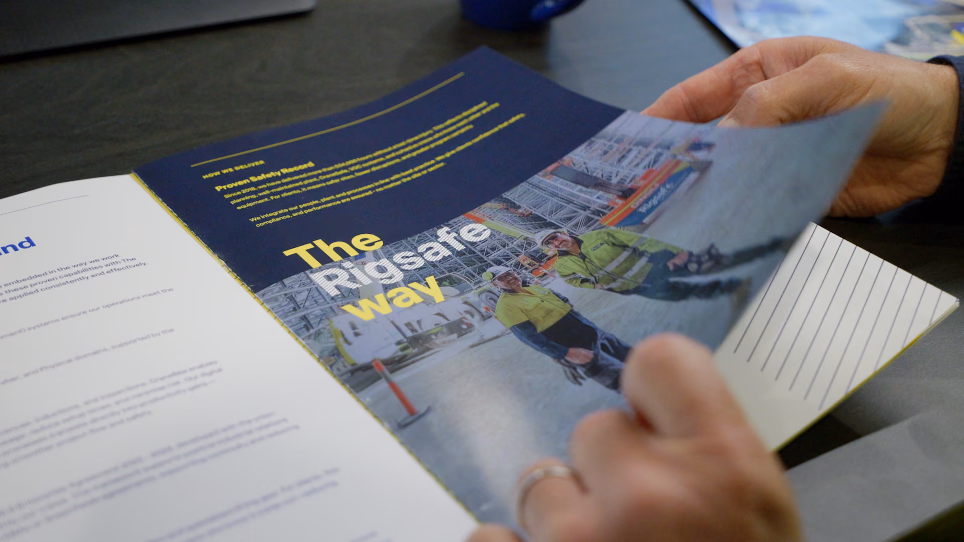 Hands flipping through a brochure page showing two workers in safety gear and text 'The Rigsafe way'.