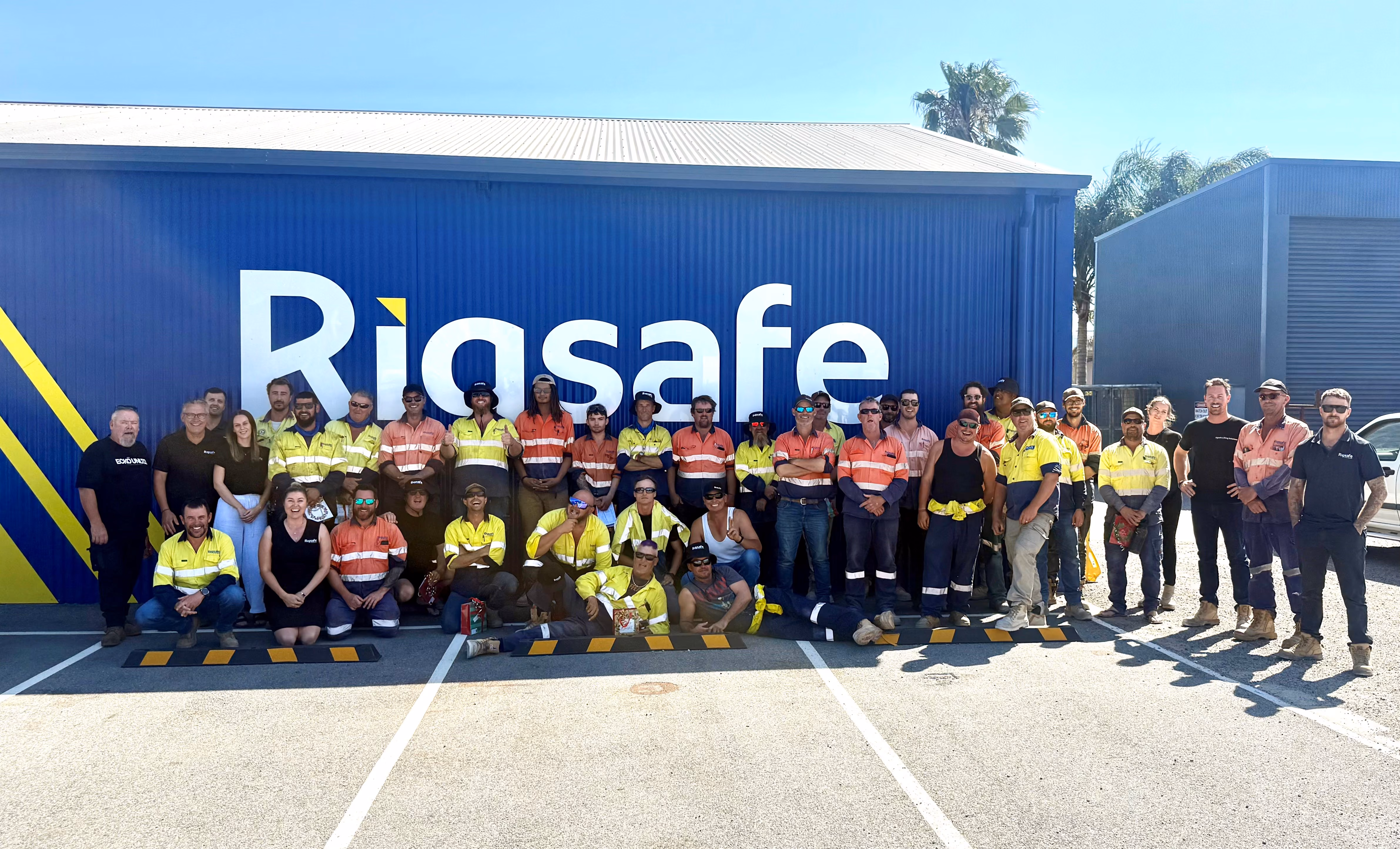 Group photo of workers in safety uniforms standing and kneeling outside a building with 'Rigasafe' sign.