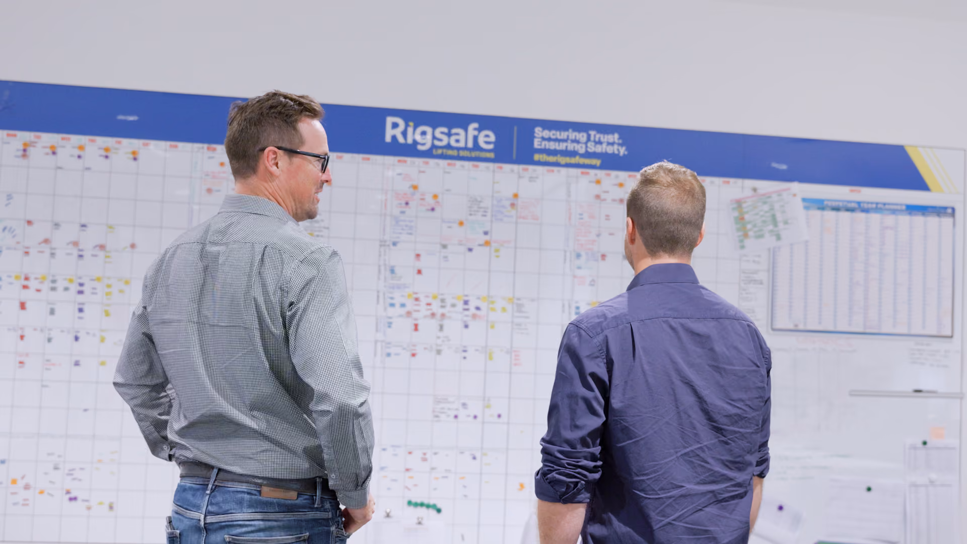 Two men standing, looking at a large Rigsafe calendar and planning board on a wall.