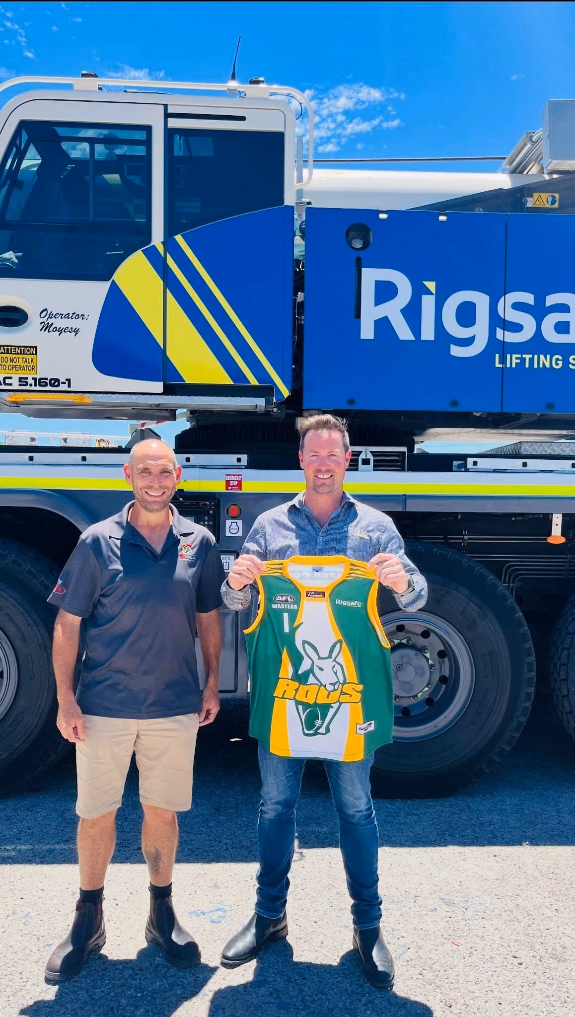 Two men stand in front of a blue RigSafe vehicle; one holds a green and yellow kangaroo sports jersey.