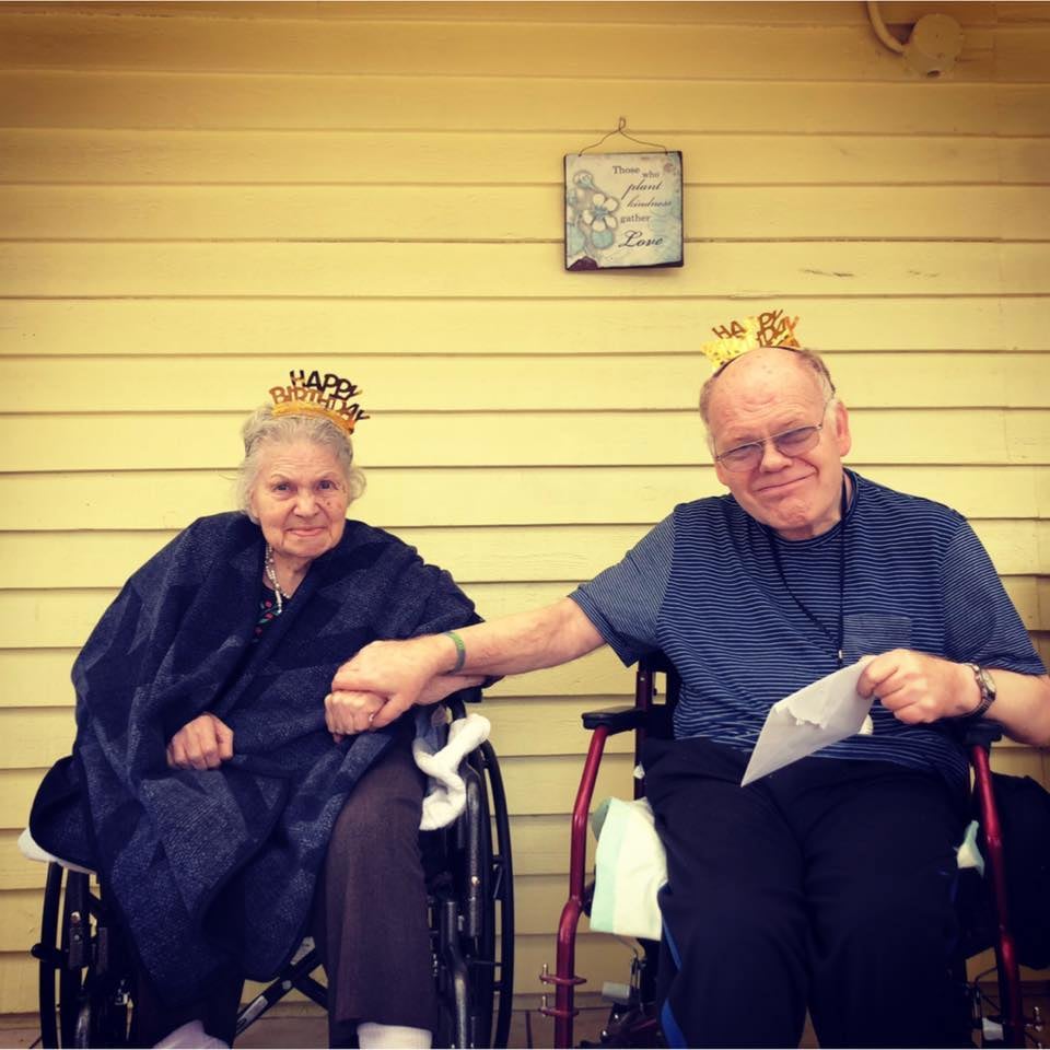 Elderly man and woman sitting in wheelchairs holding hands, both wearing 'Happy Birthday' headbands, with a wooden yellow wall and a framed floral sign behind them.