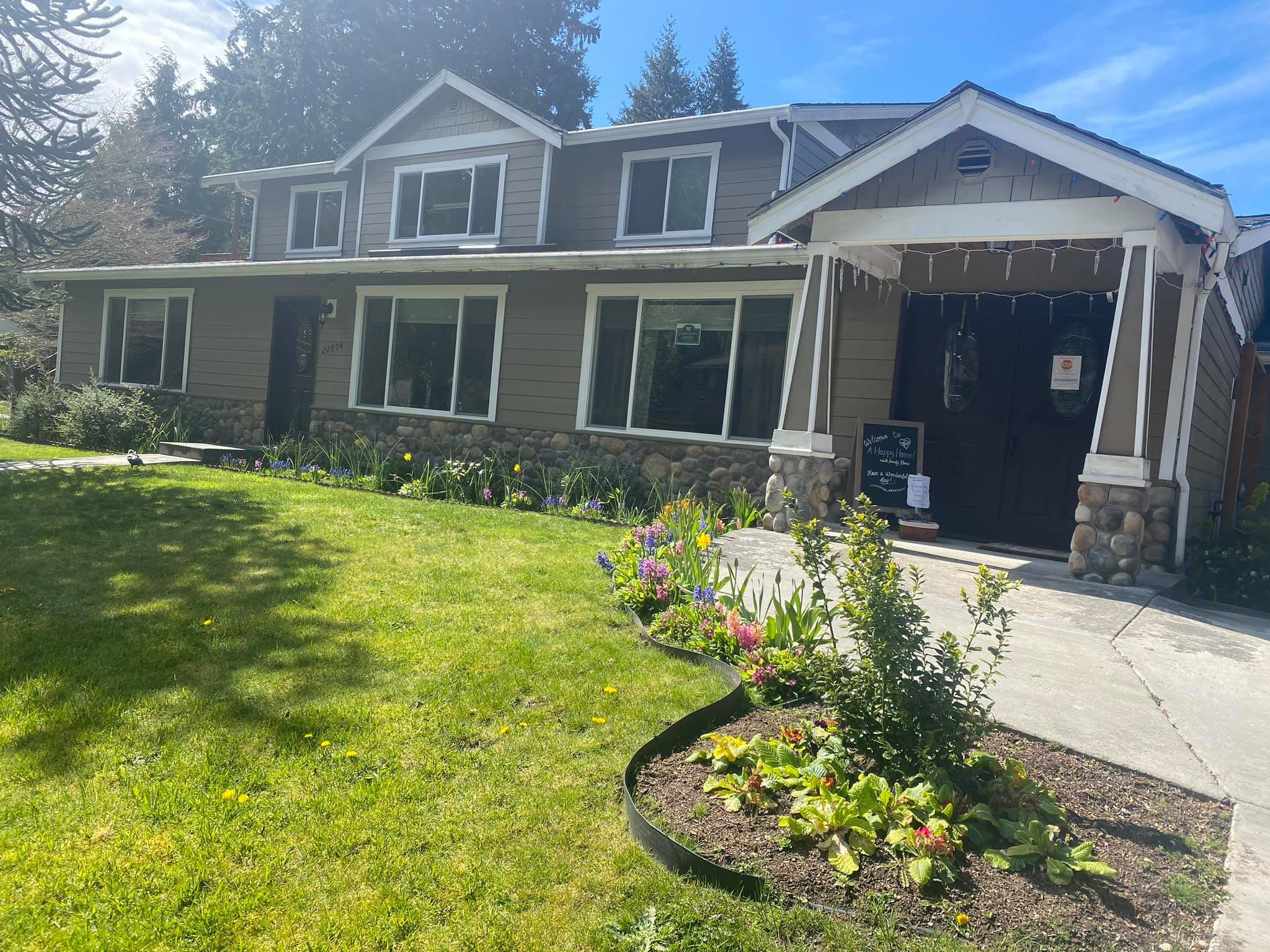 Two-story gray house with stone foundation, dark double front doors, a flower garden border, and a green lawn under a clear blue sky.