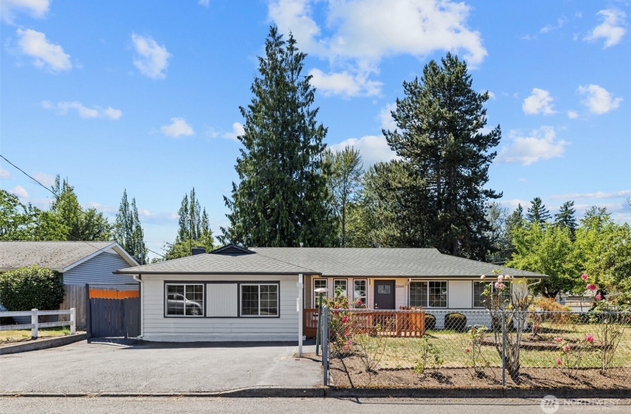 Single-story white house with gray roof, large windows, and a fenced front yard with rose bushes under a blue sky with scattered clouds.