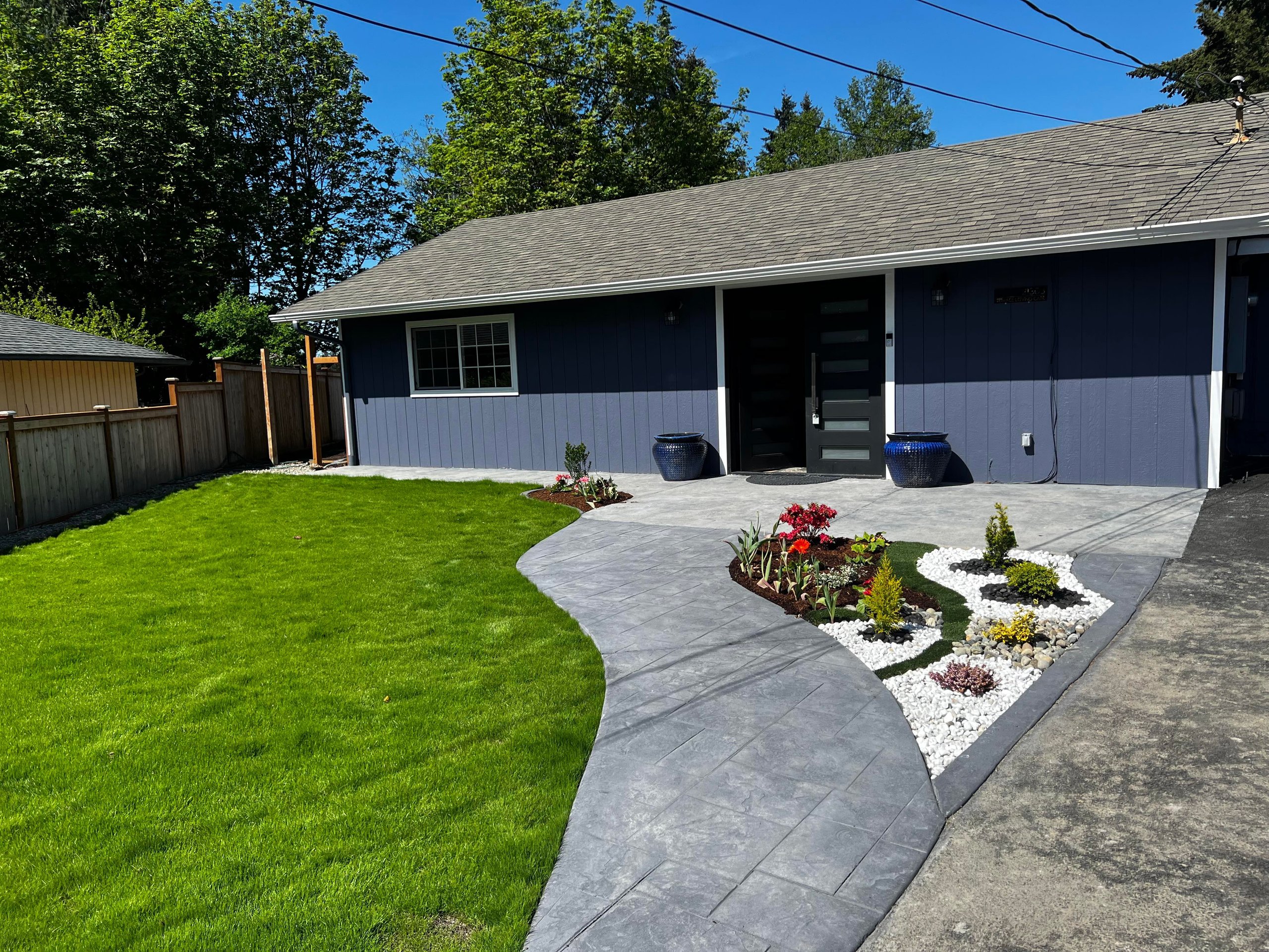 Front yard of a gray house with a curved concrete walkway, green lawn, and decorative flower beds with white rocks.