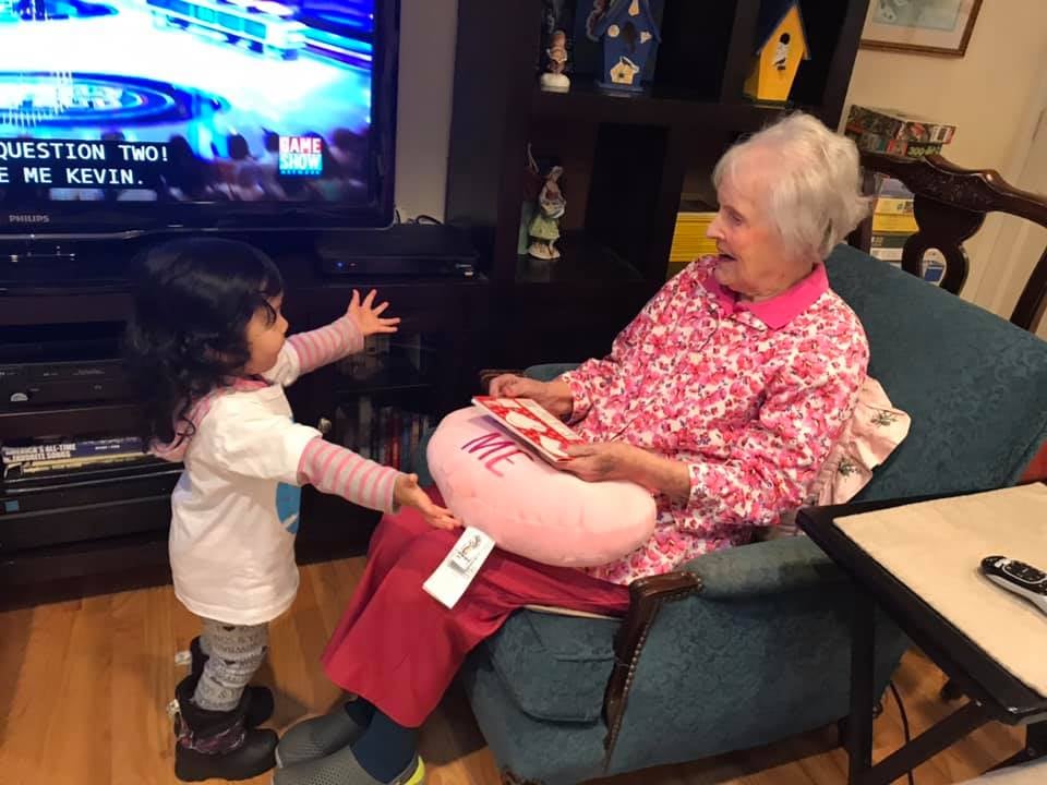Toddler with arms outstretched towards a seated elderly woman holding a heart-shaped pillow in a living room.