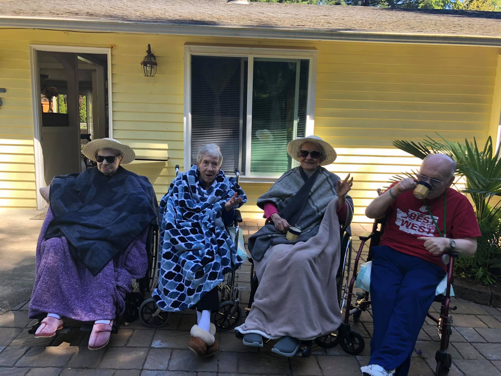 Four elderly people sitting outdoors in wheelchairs and a walker, wrapped in blankets and wearing hats and sunglasses, with a yellow house in the background.