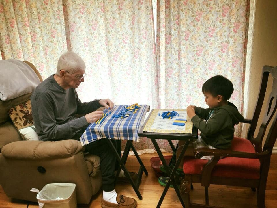 An elderly man and a young boy sitting across from each other at small tables, playing with blue and yellow building blocks indoors.