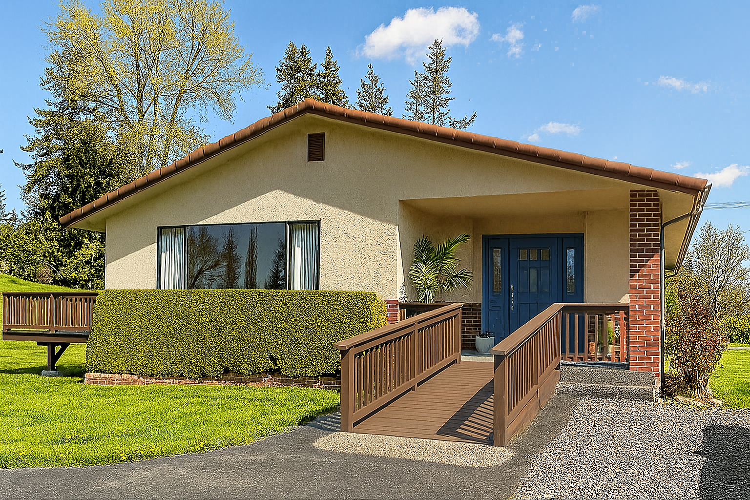 Single-story house with beige stucco, blue front door, brick accents, wooden ramp, and neatly trimmed hedge under a clear sky.