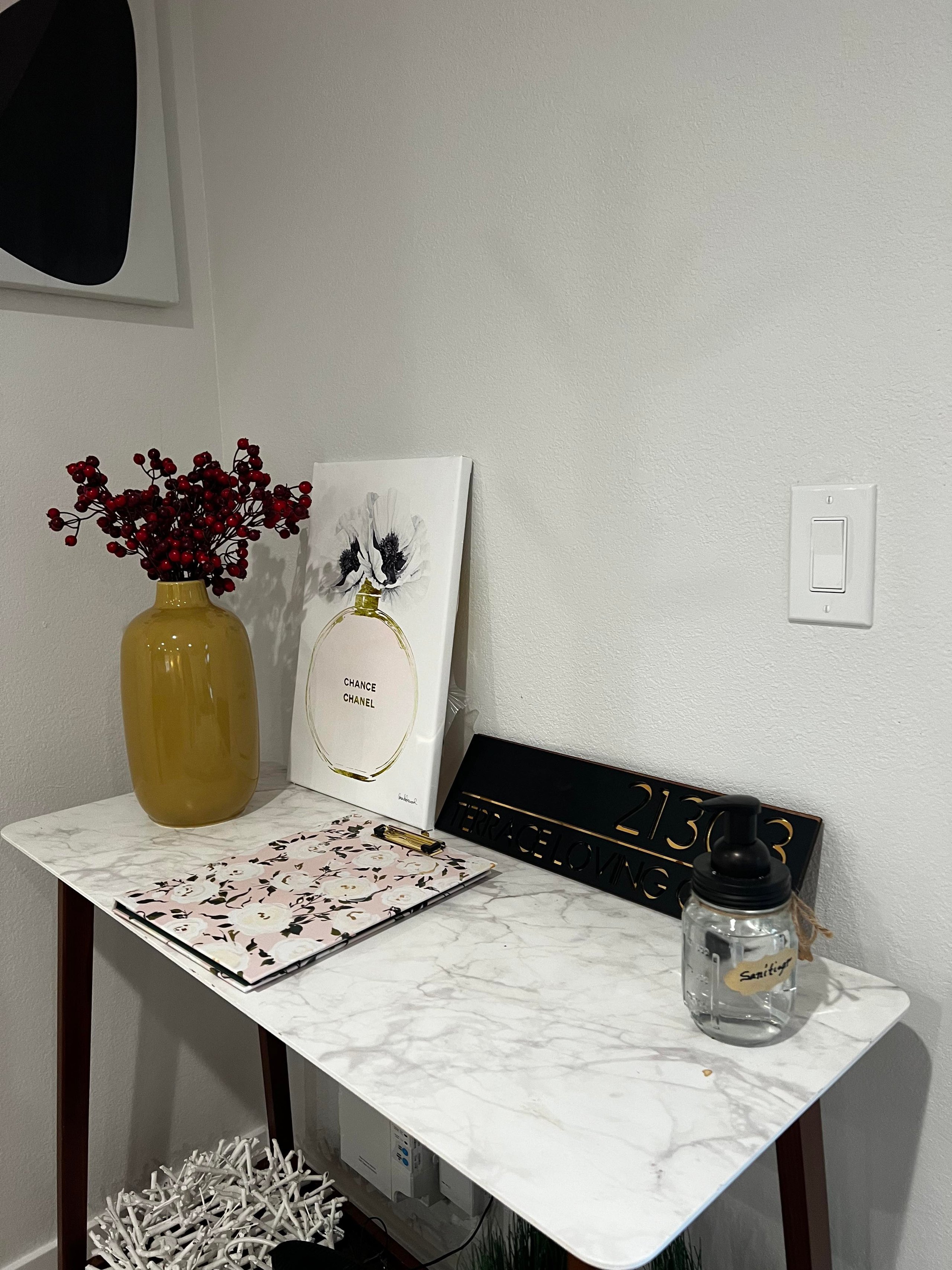 Marble-top table with a mustard yellow vase holding red berries, a floral clipboard, a framed Chanel Chance perfume print, a black sign with gold lettering, and a clear hand sanitizer bottle.