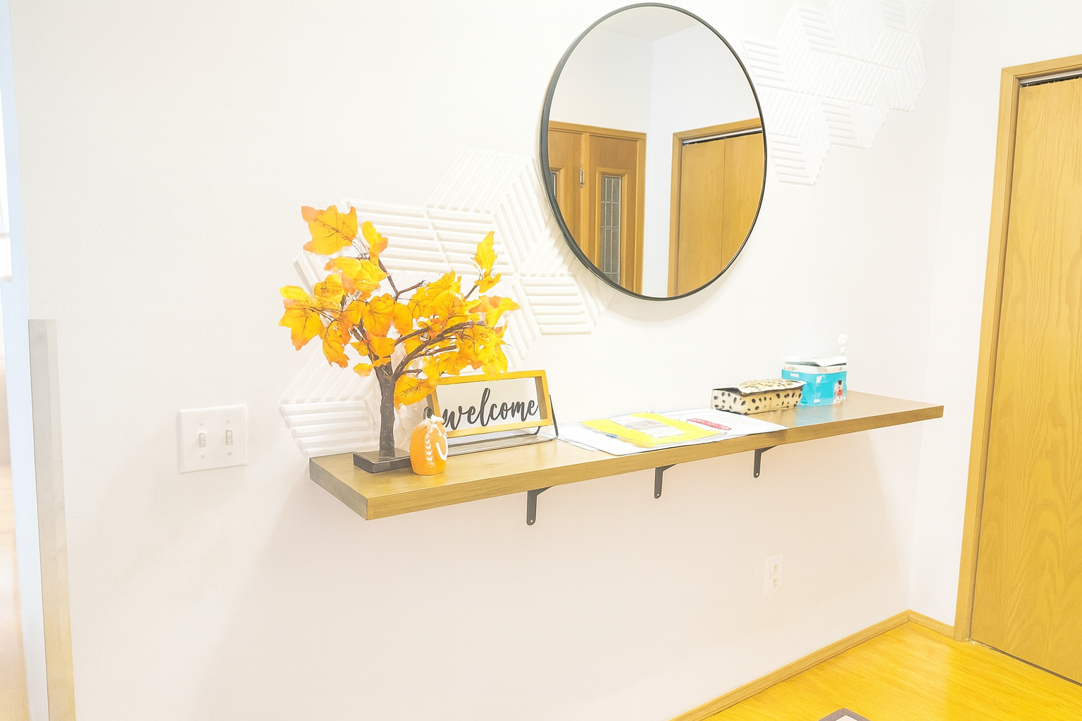 Wall-mounted wooden shelf with autumn leaves decor, a welcome sign, a small pumpkin, and tissues under a round mirror.
