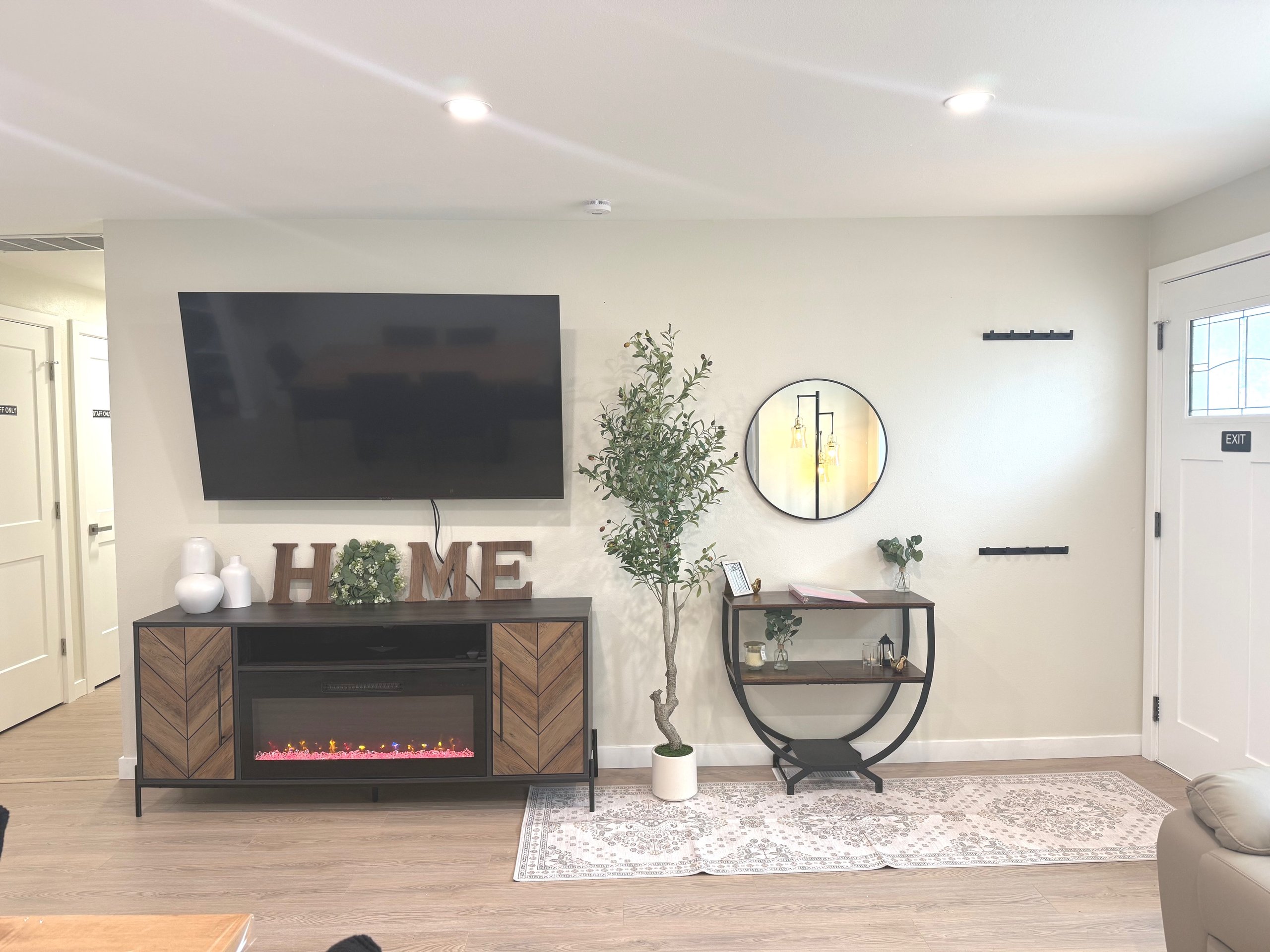 Modern living room wall with a mounted flat-screen TV above a wooden console with an electric fireplace and decorative HOME letters, next to a tall potted plant and a round mirror above a small black and wood shelf.