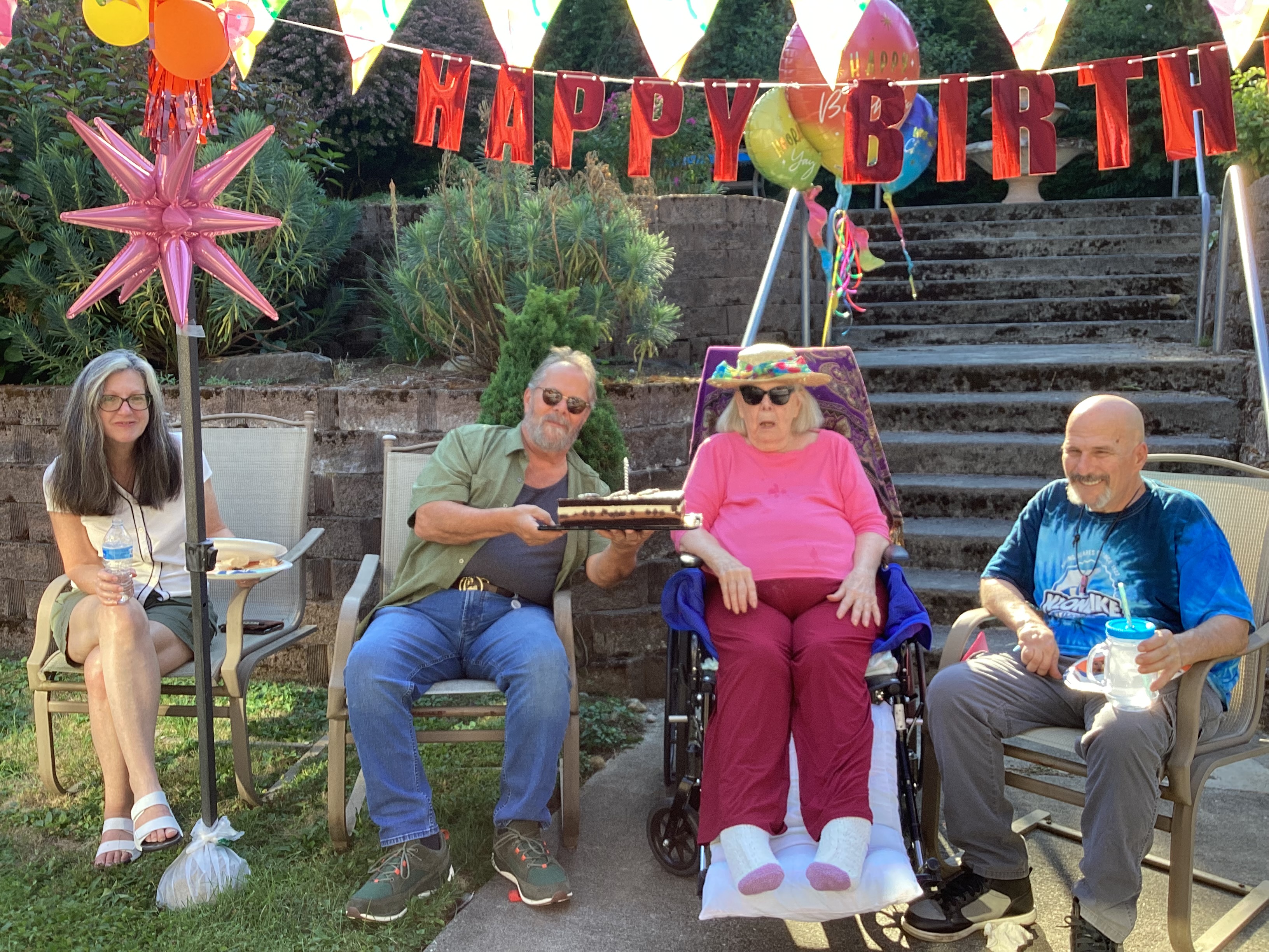 Four people seated outdoors with birthday decorations, including a banner and balloons; a man holds a birthday cake with a candle.