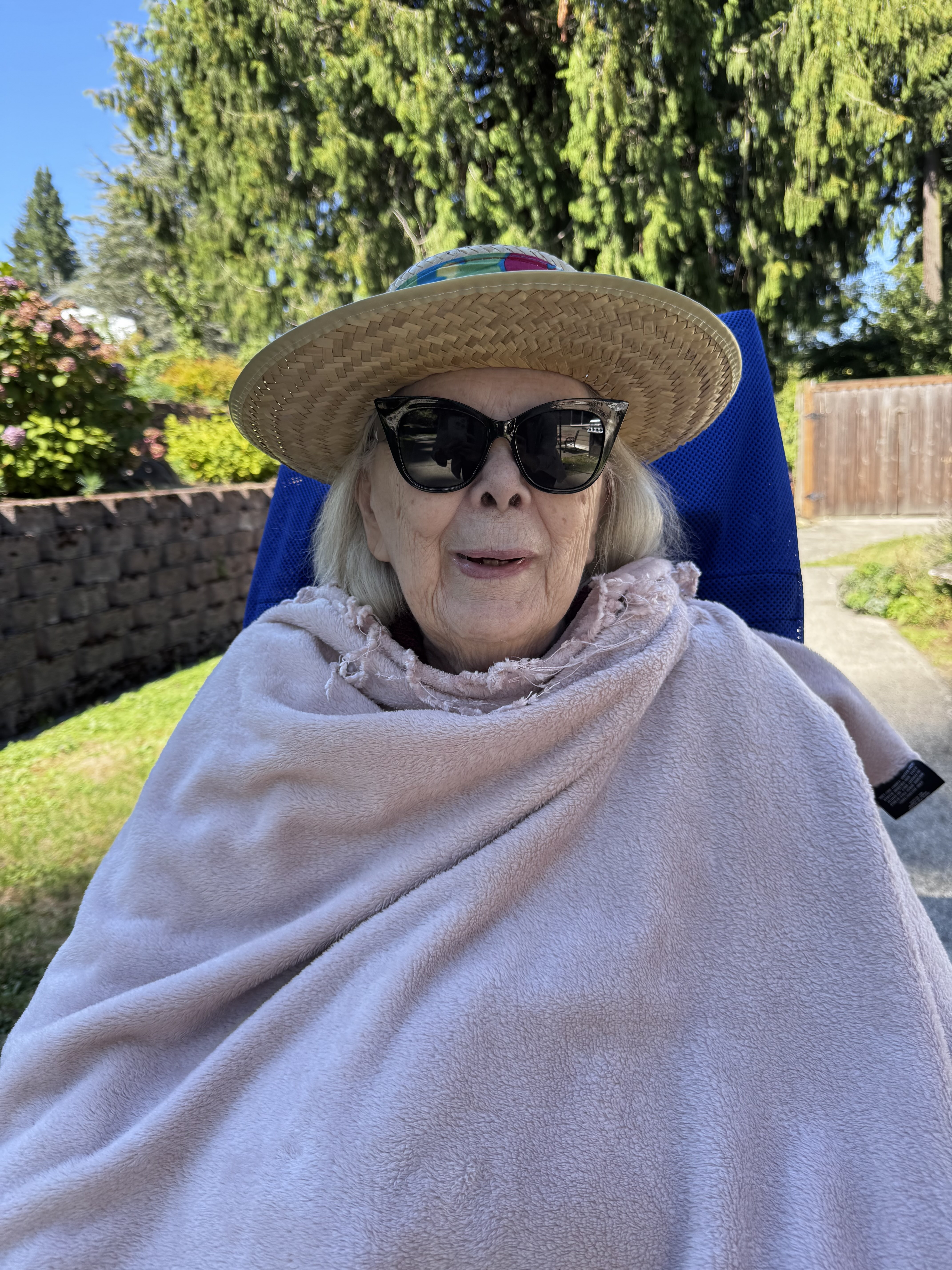 Elderly woman wearing a straw hat and dark sunglasses, wrapped in a light pink blanket, sitting outdoors in a blue chair.