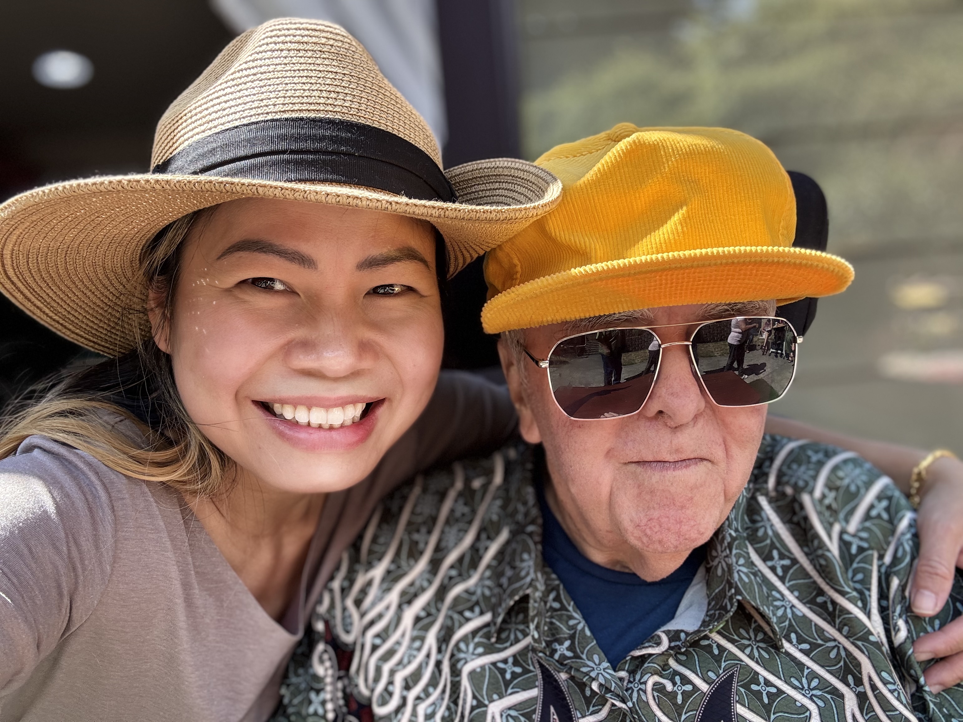 Smiling woman wearing a straw hat poses closely with an elderly man wearing a bright yellow cap and sunglasses.