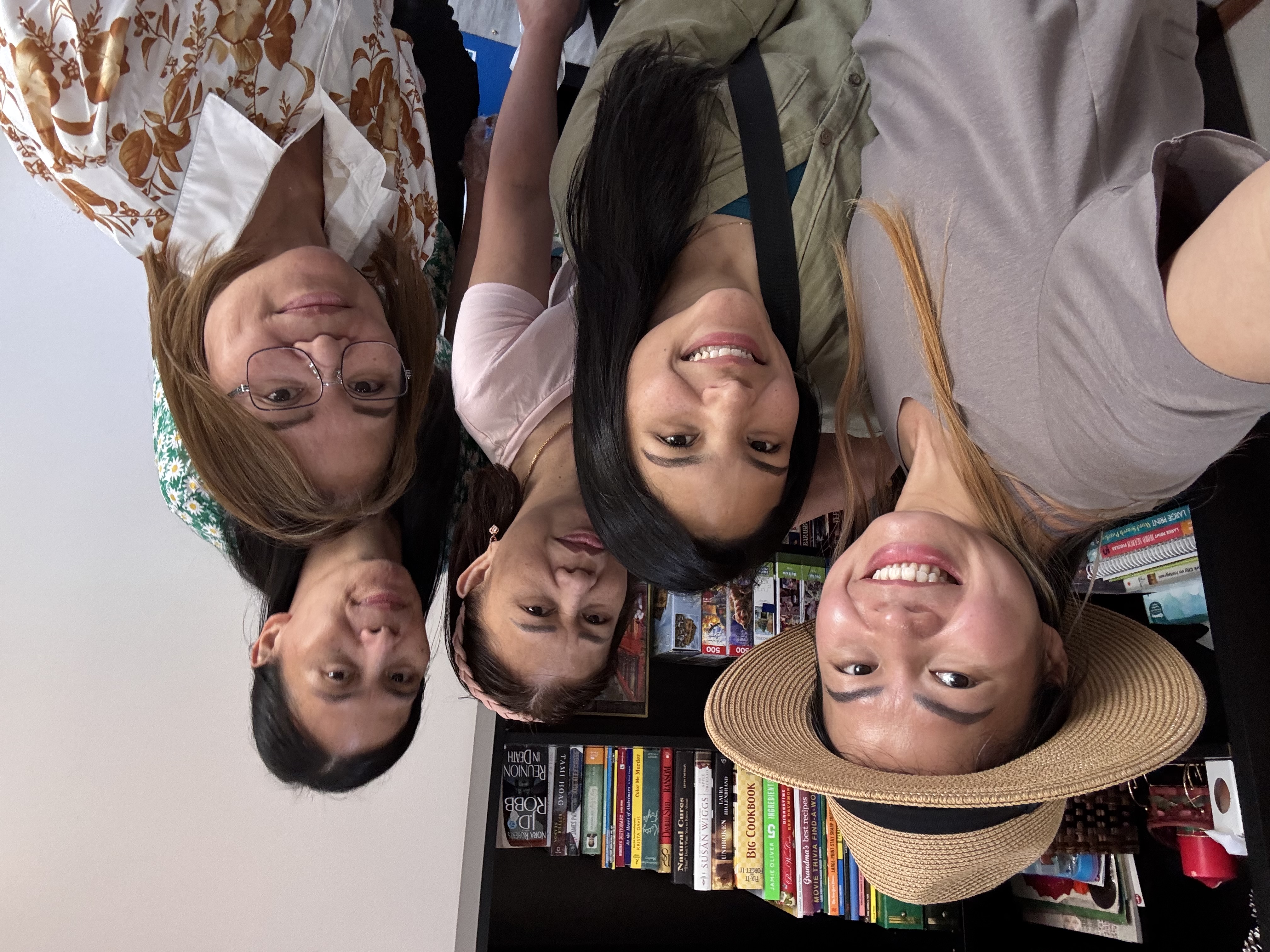 Group of five smiling women taking a selfie indoors with a bookshelf filled with books and puzzles in the background.