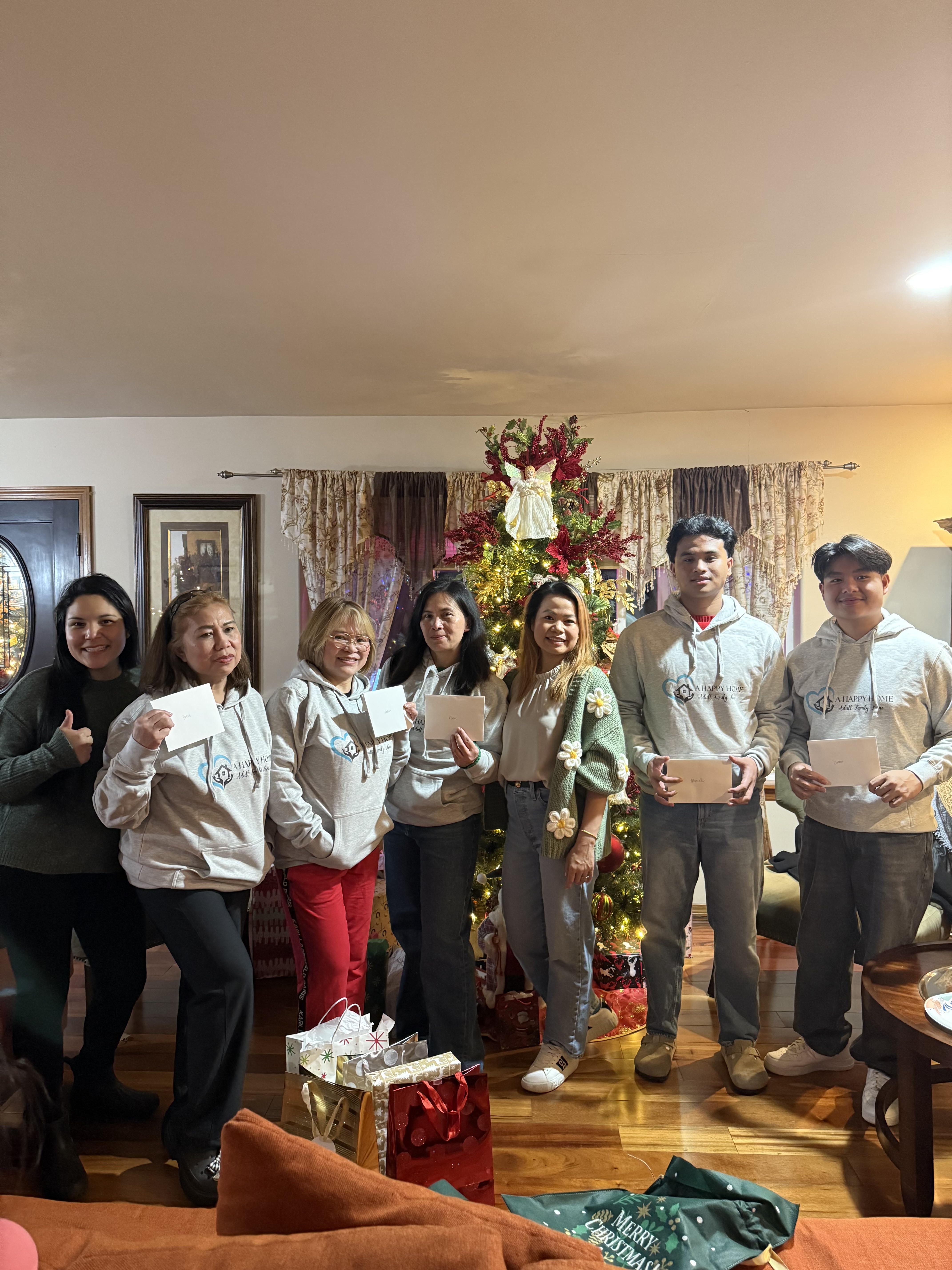 Seven people standing indoors in front of a decorated Christmas tree holding envelopes and gift bags on the floor.
