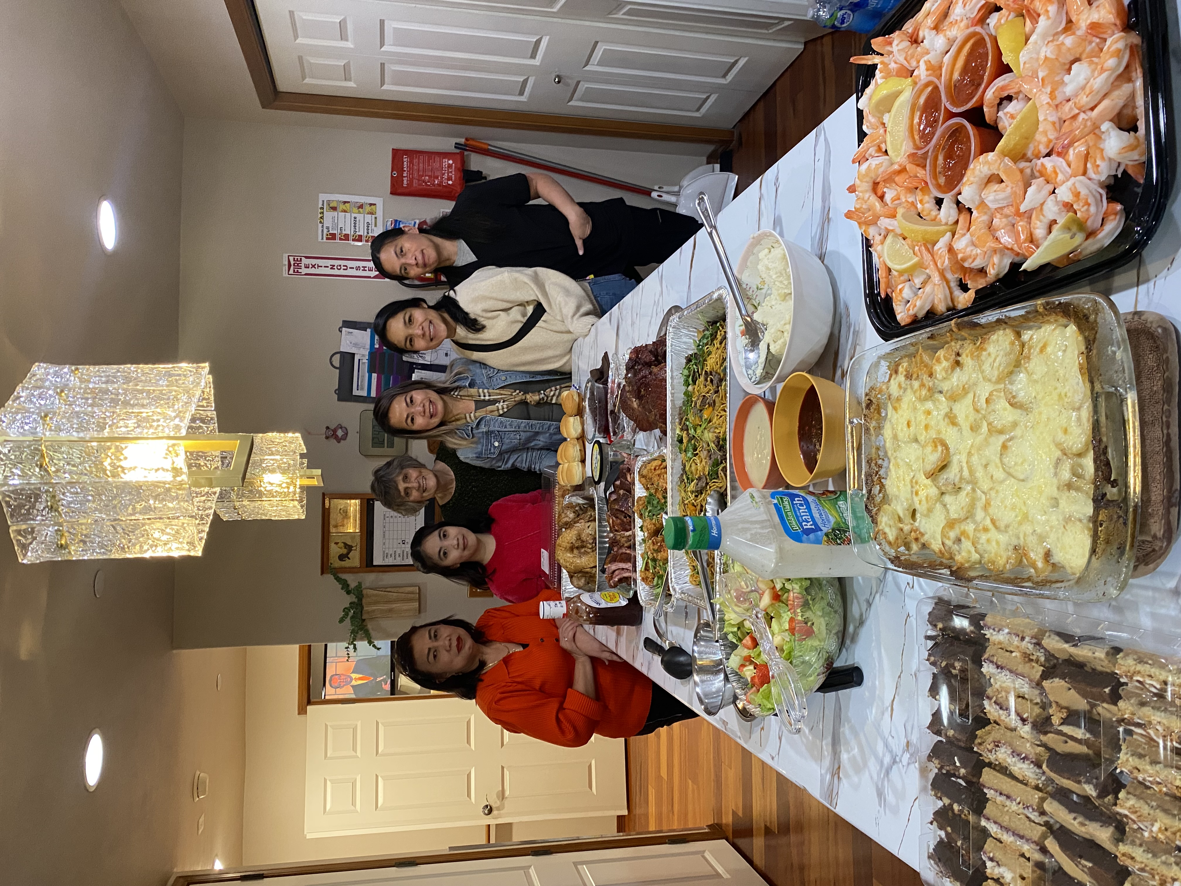 Six women standing behind a table filled with various dishes including shrimp cocktail, scalloped potatoes, salad, ribs, rolls, and sauces in a dining room.
