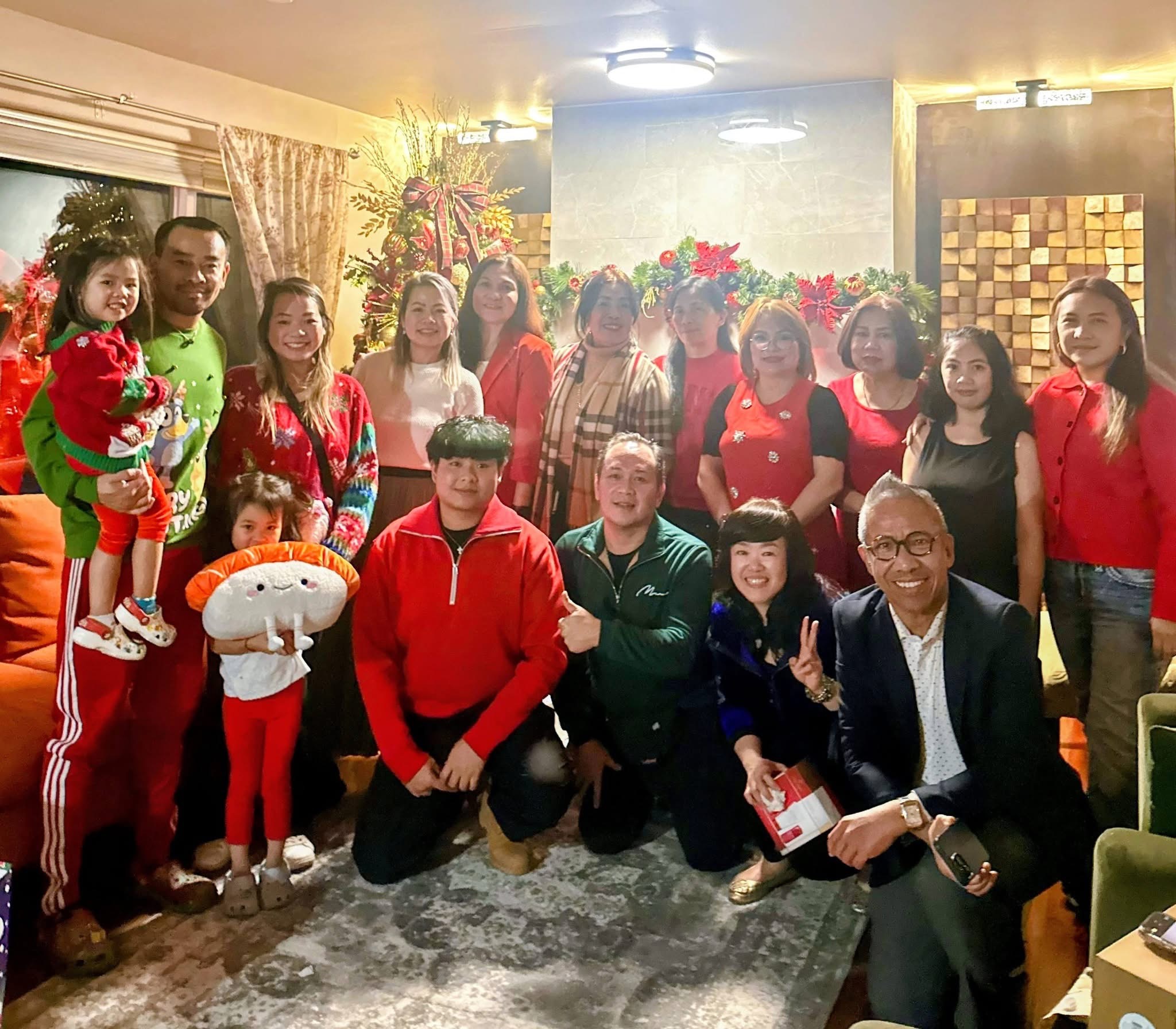 Group of adults and children posing indoors in festive holiday clothes with Christmas decorations in the background.