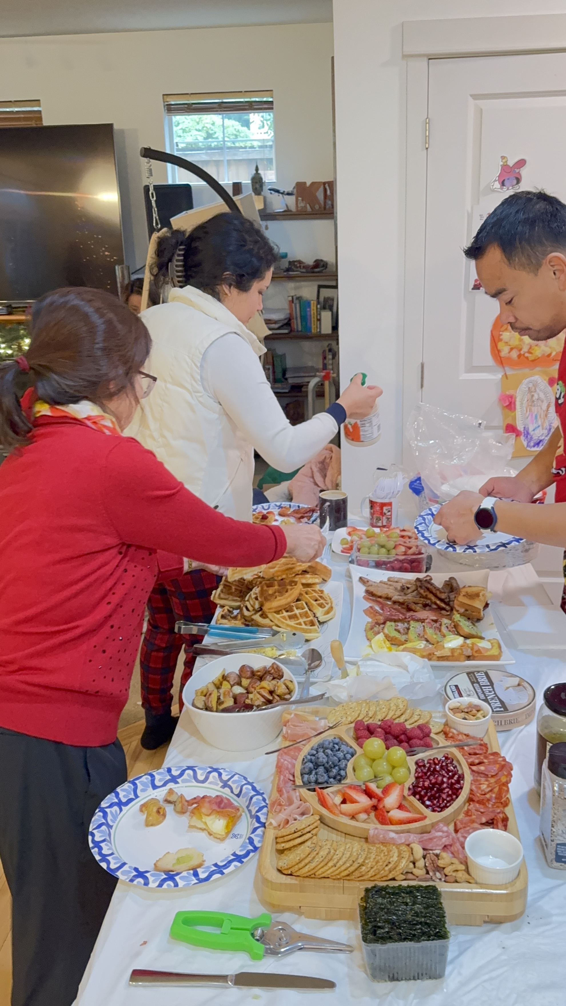 Three people serving themselves food from a table with waffles, fruits, crackers, meats, and various dishes at a casual indoor gathering.