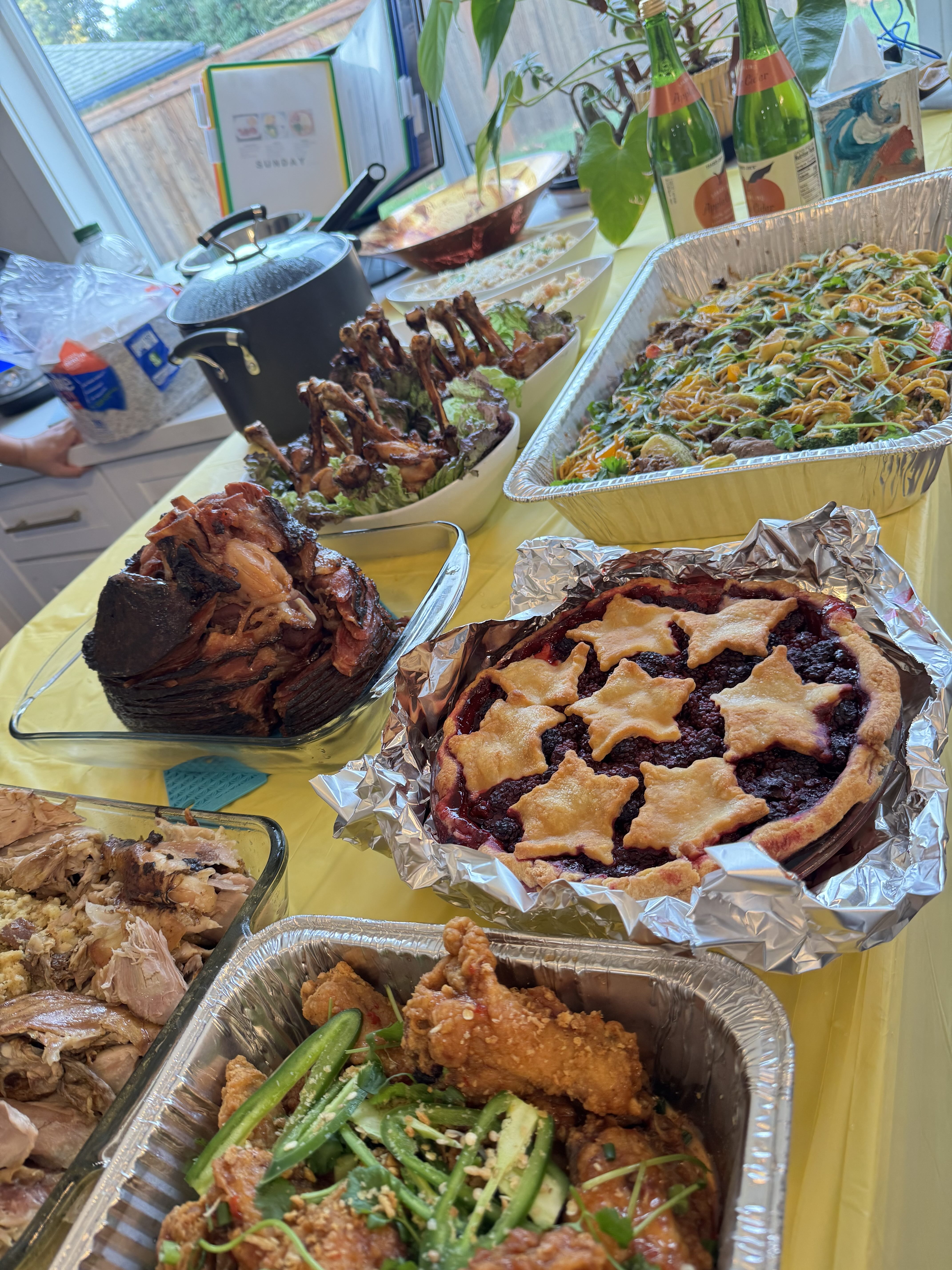 Table with a variety of homemade dishes including a berry pie with star-shaped crust pieces, fried chicken with sliced jalapeños, sliced ham, chicken wings, noodles with vegetables, and two bottles of apple cider.