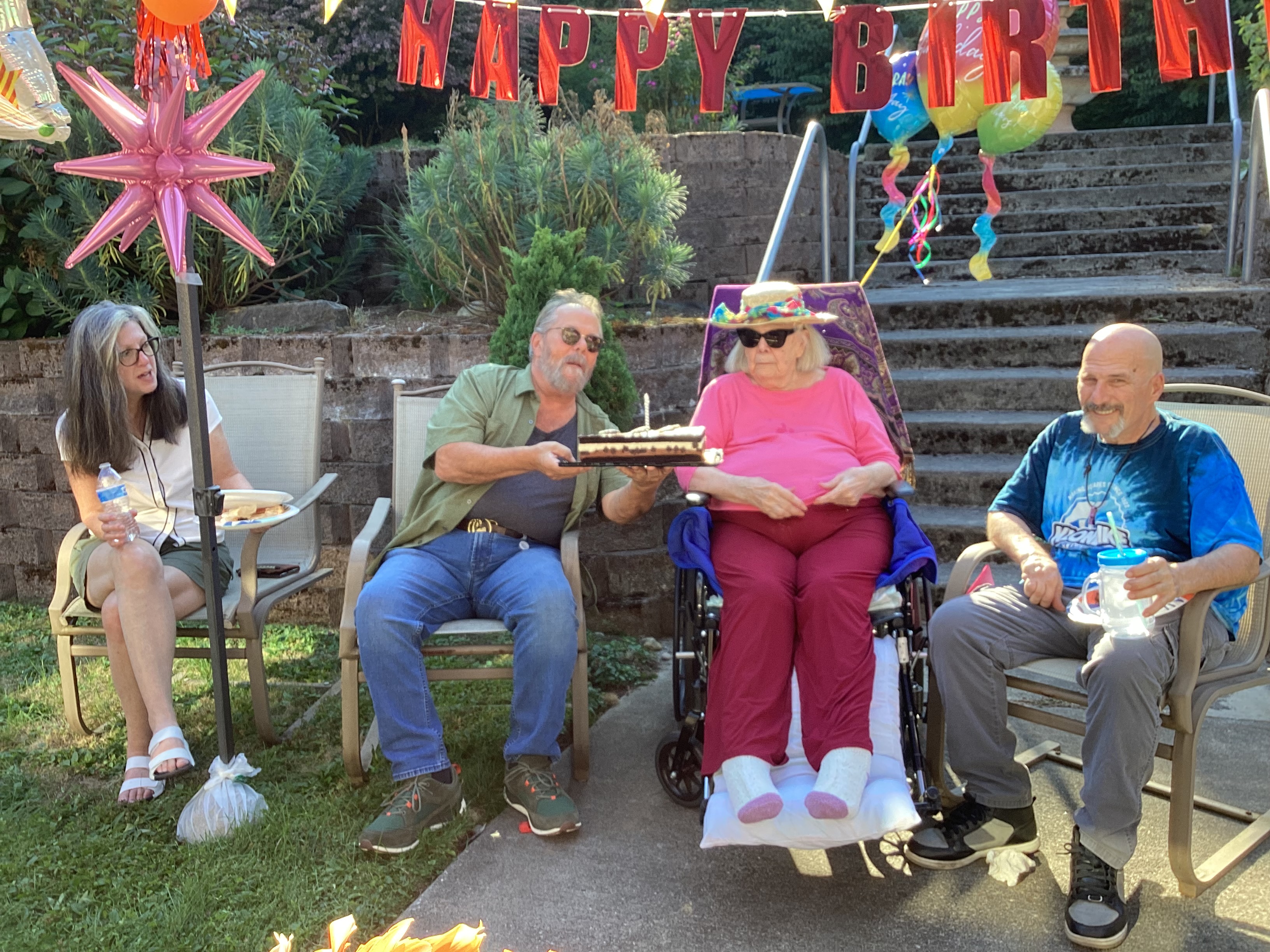 Four people seated outdoors at a birthday party, with a man holding a cake with lit candles near an elderly woman in a wheelchair wearing a hat and sunglasses, and a 'Happy Birthday' banner overhead.
