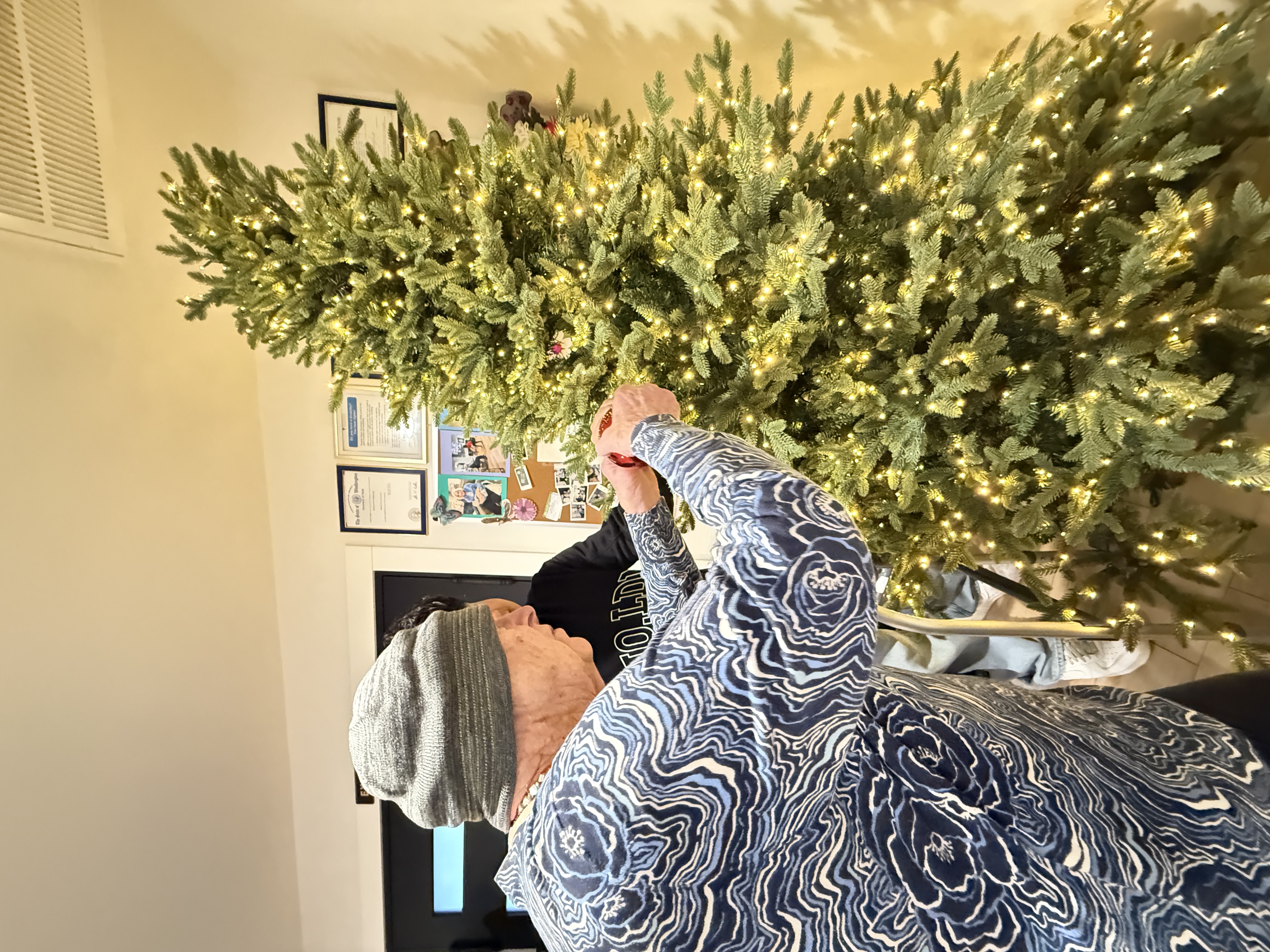Two people decorating a lit artificial Christmas tree indoors.
