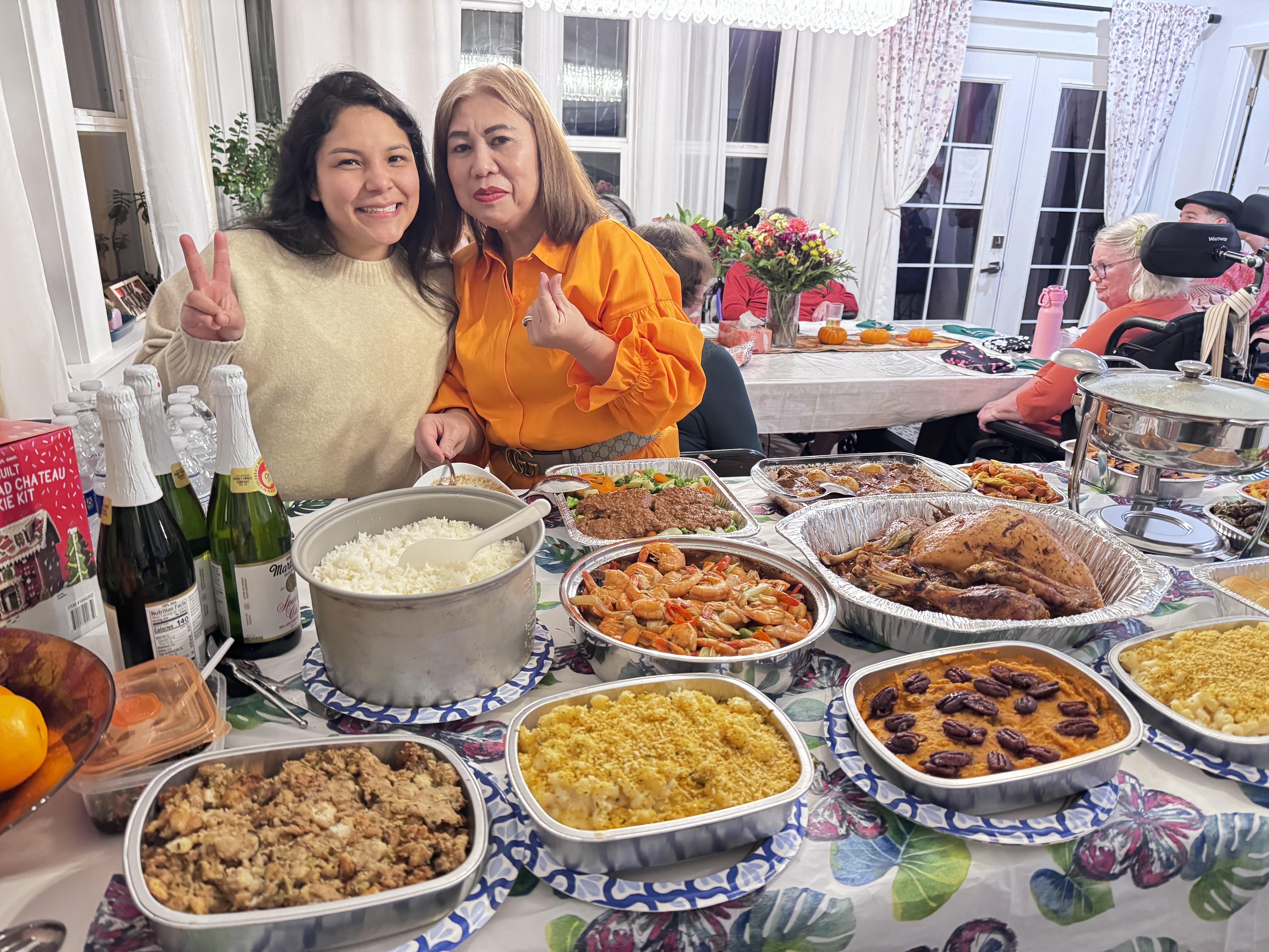 Two women smiling and posing with peace and heart fingers near a table full of various cooked dishes and drinks at a family gathering.