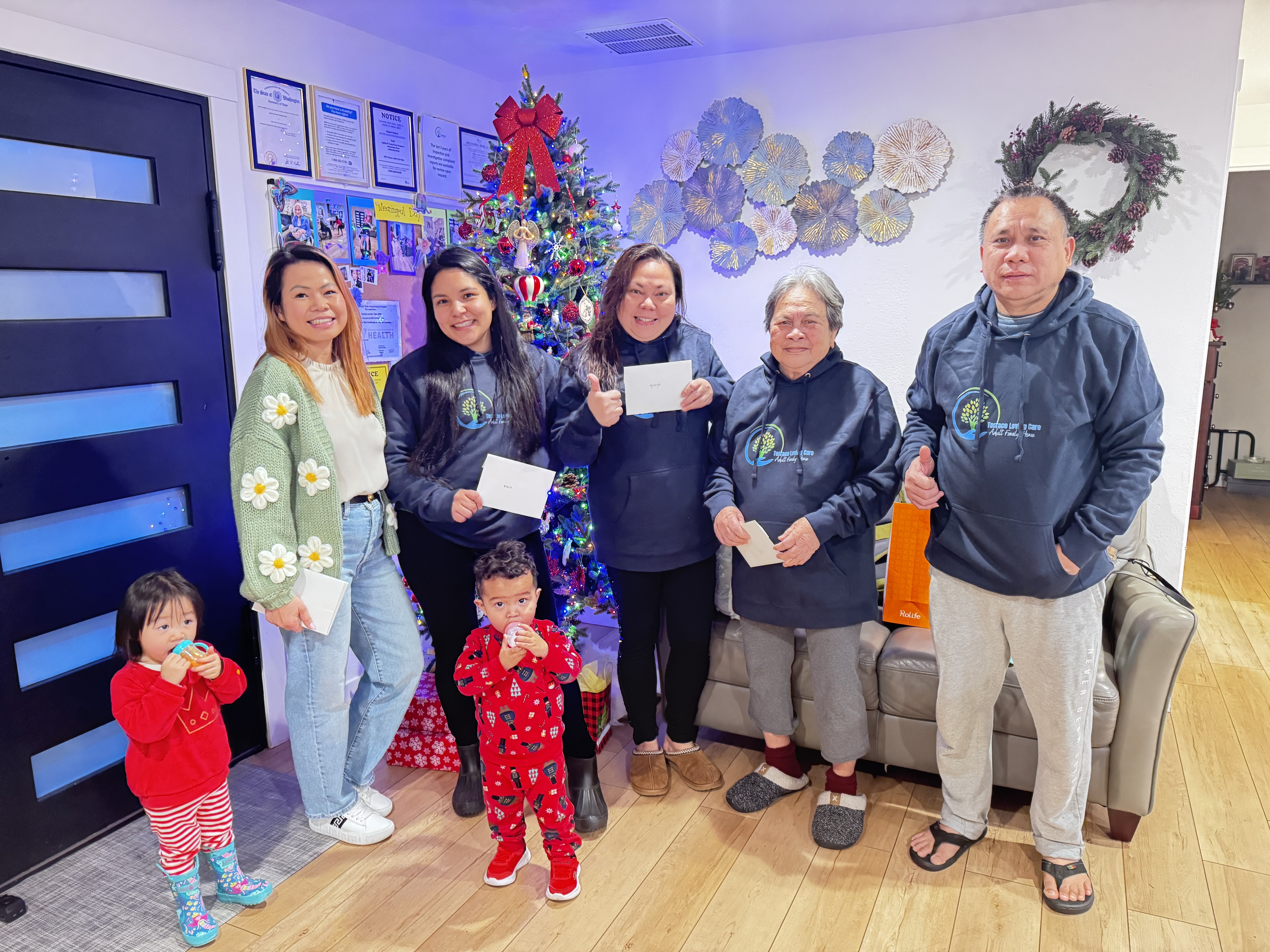 Six people including two children standing indoors in front of a decorated Christmas tree and holiday wreaths, some holding envelopes.