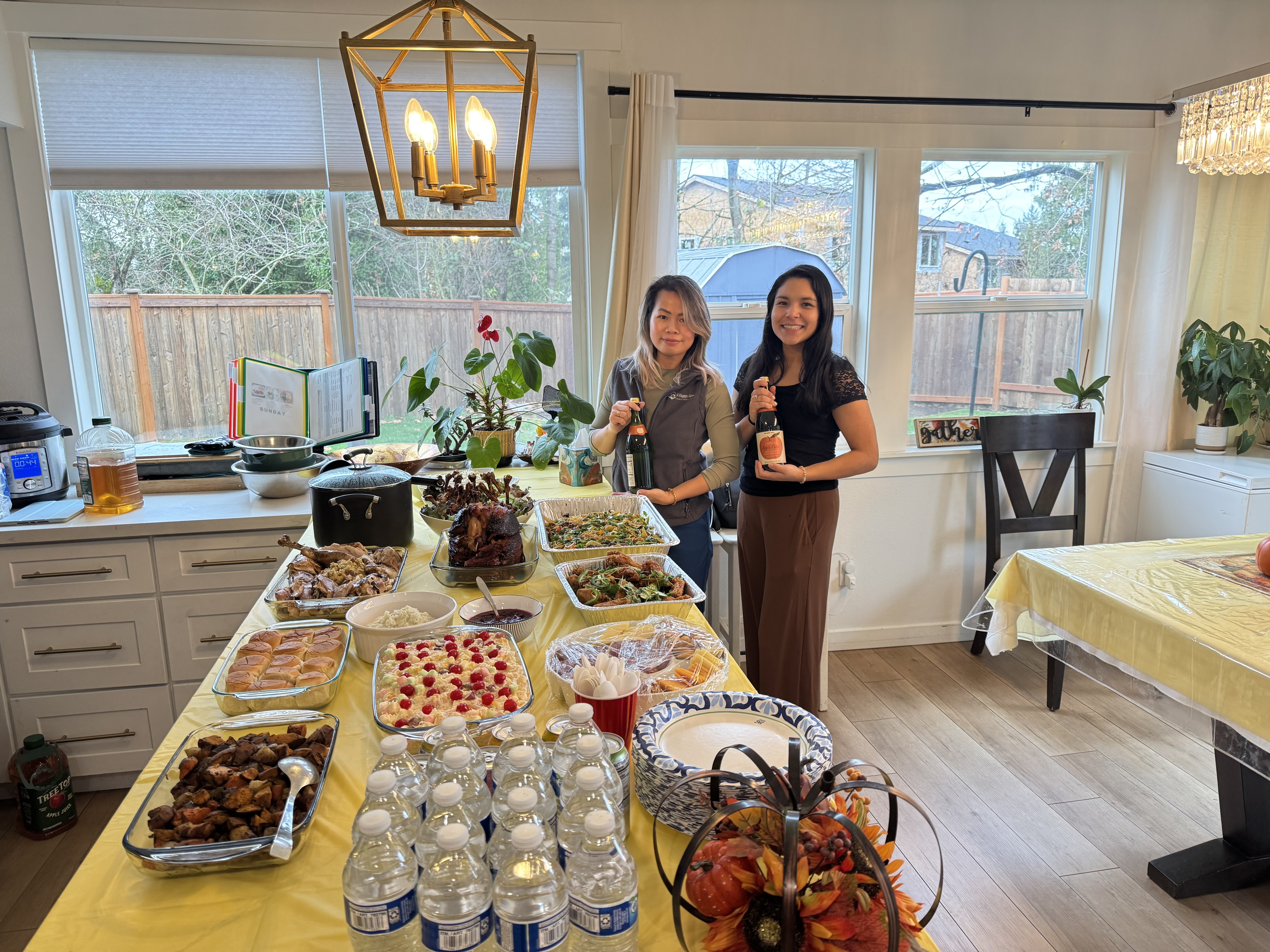 Two women standing behind a long table filled with home-cooked Thanksgiving food dishes and bottled water in a dining room.