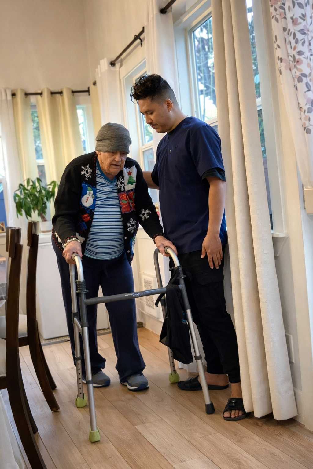Caregiver assisting elderly man with walker inside a home near windows with curtains.