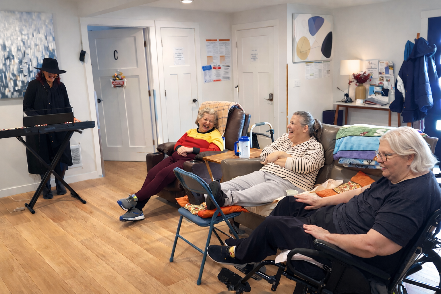 Woman playing keyboard and smiling as three elderly women seated and laughing in a cozy room.
