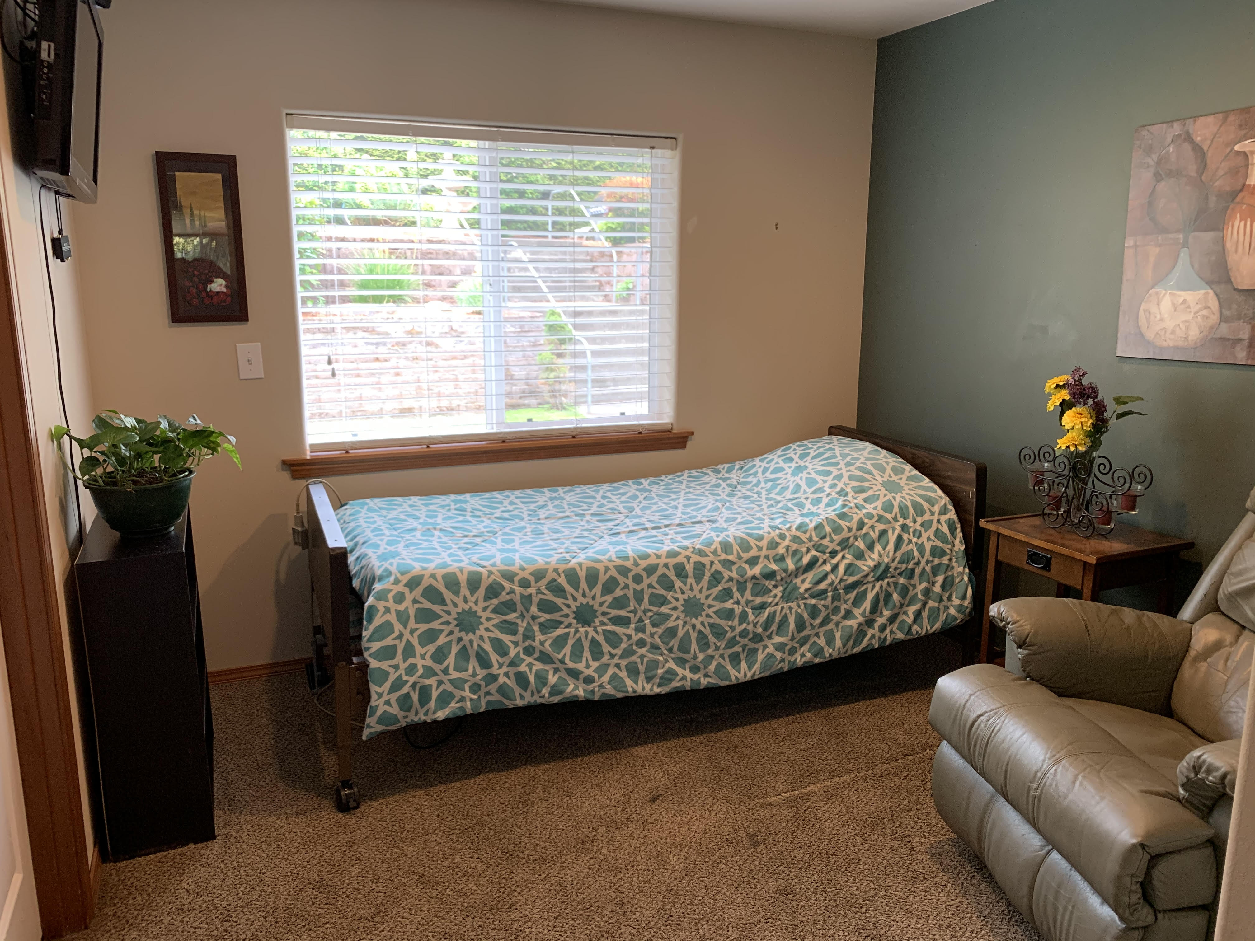 Small bedroom with a single bed covered by a blue and white patterned blanket, a beige recliner chair, a window with blinds, a wooden nightstand with a flower vase, and a green accent wall.