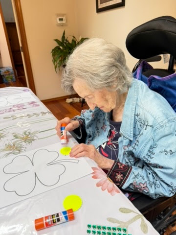 Elderly woman with gray hair sitting in a wheelchair, using glue stick on paper with clover outlines for a craft activity.