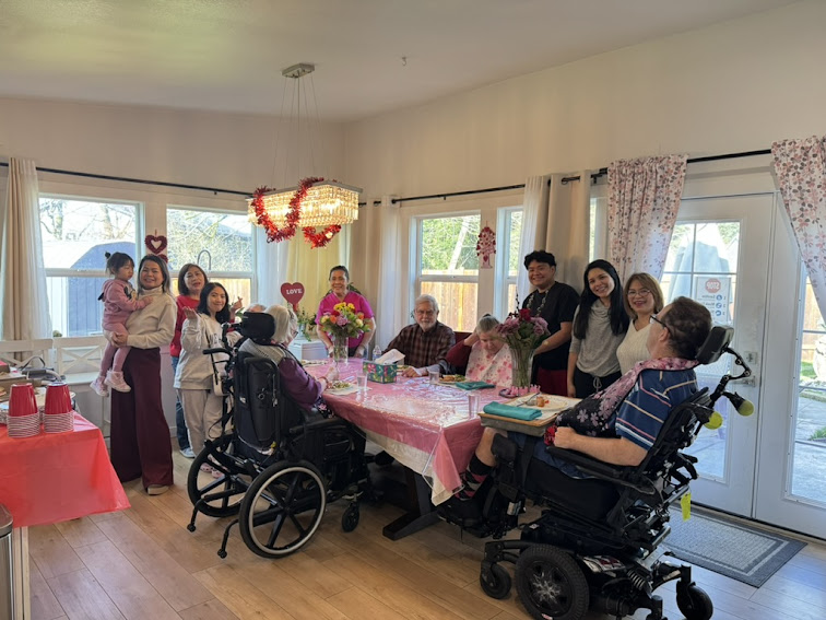Multigenerational group of people gathered around a dining table decorated with flowers and Valentine’s Day themed decorations in a bright room.