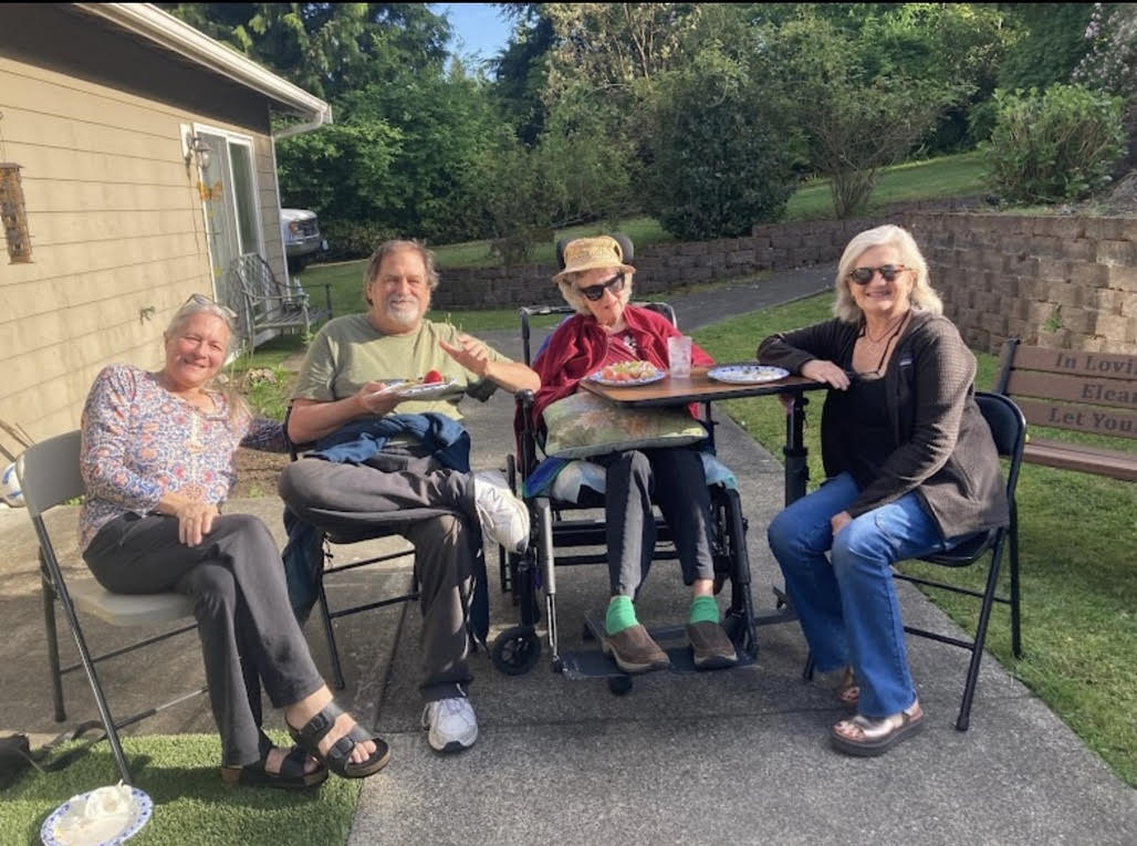 Four adults sitting outdoors on chairs and a wheelchair, enjoying food and smiling in a sunny backyard.