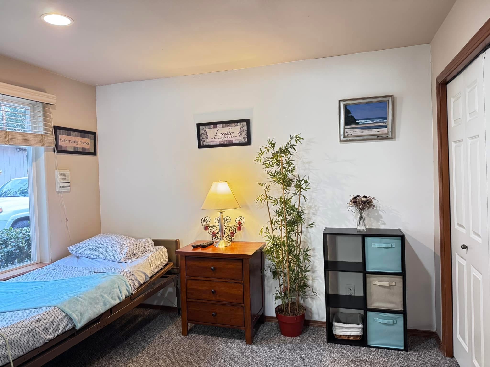 Small bedroom with a bed next to a window, a wooden nightstand with a lamp, a potted plant, and a black cubby shelf with storage bins and a vase of flowers.