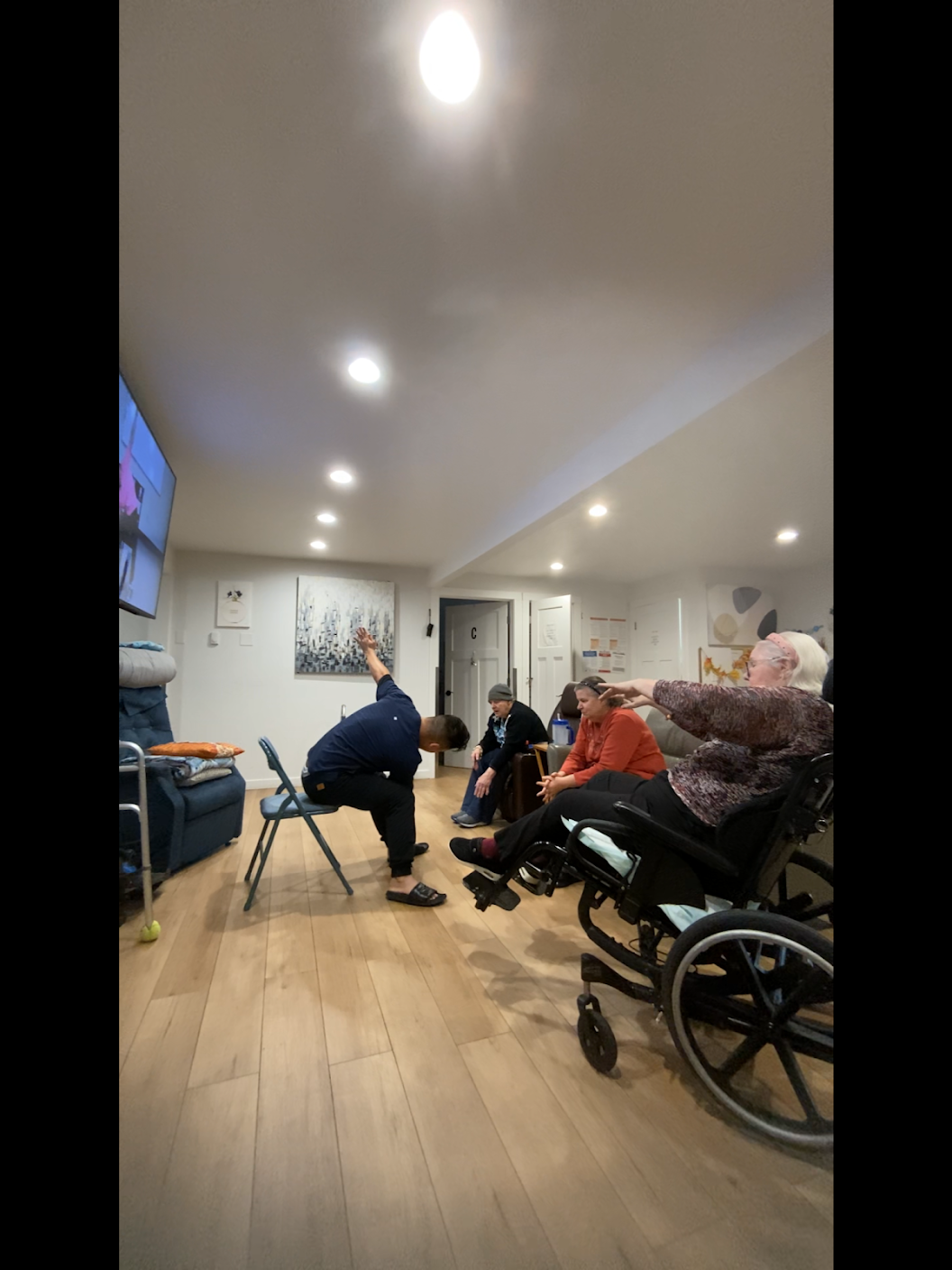 A group of three people seated indoors, with one person in a wheelchair and another performing a seated stretch exercise.