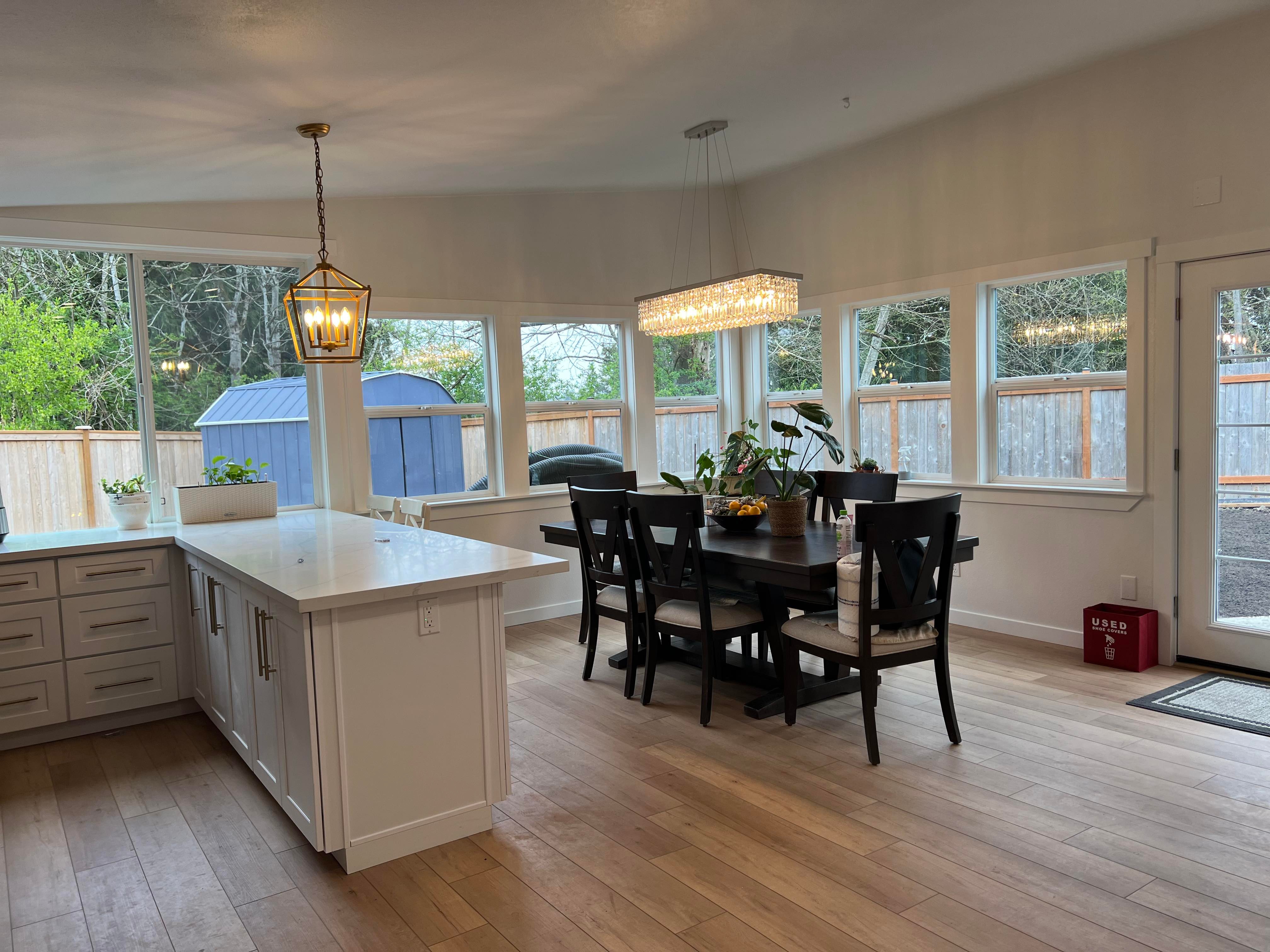 Modern kitchen dining area with wooden floor, dark wood table surrounded by six chairs, large windows, white cabinets, and two pendant lights.