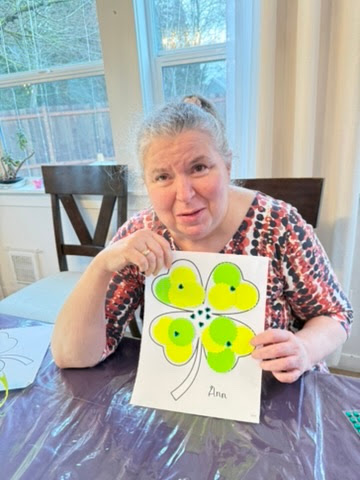 Woman sitting at a table holding a paper with a large four-leaf clover drawing colored in green and yellow paint, signed 'Ann'.