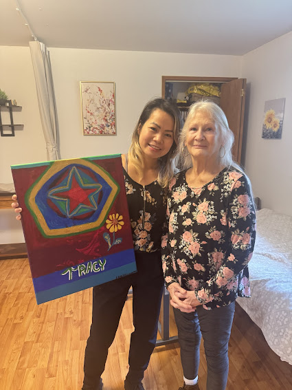 Two women standing indoors, one holding a colorful painting with a star and flower design and the name 'Tracy' at the bottom.