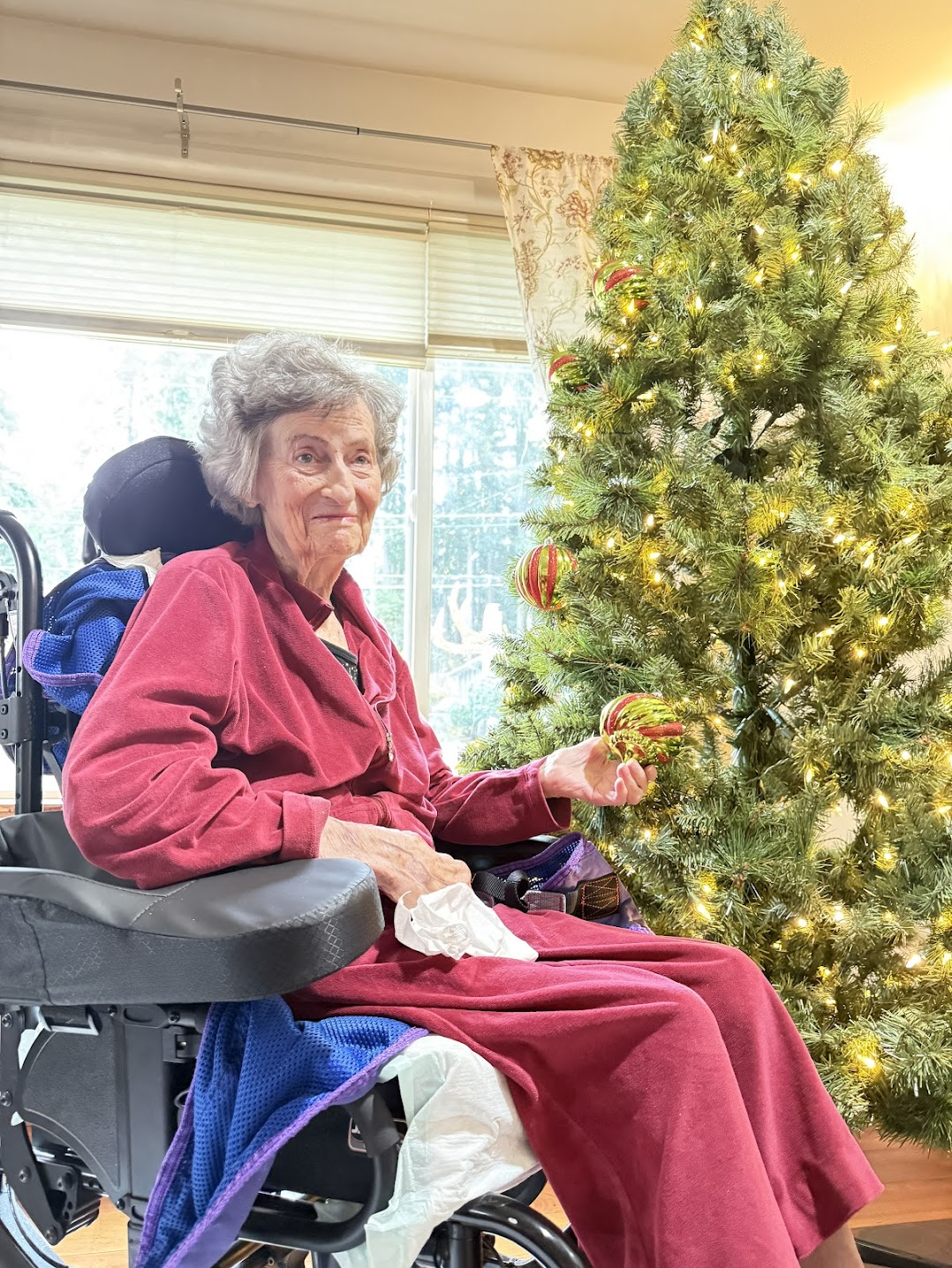 Elderly woman in a wheelchair wearing a red robe, holding a Christmas ornament next to a lit decorated Christmas tree.
