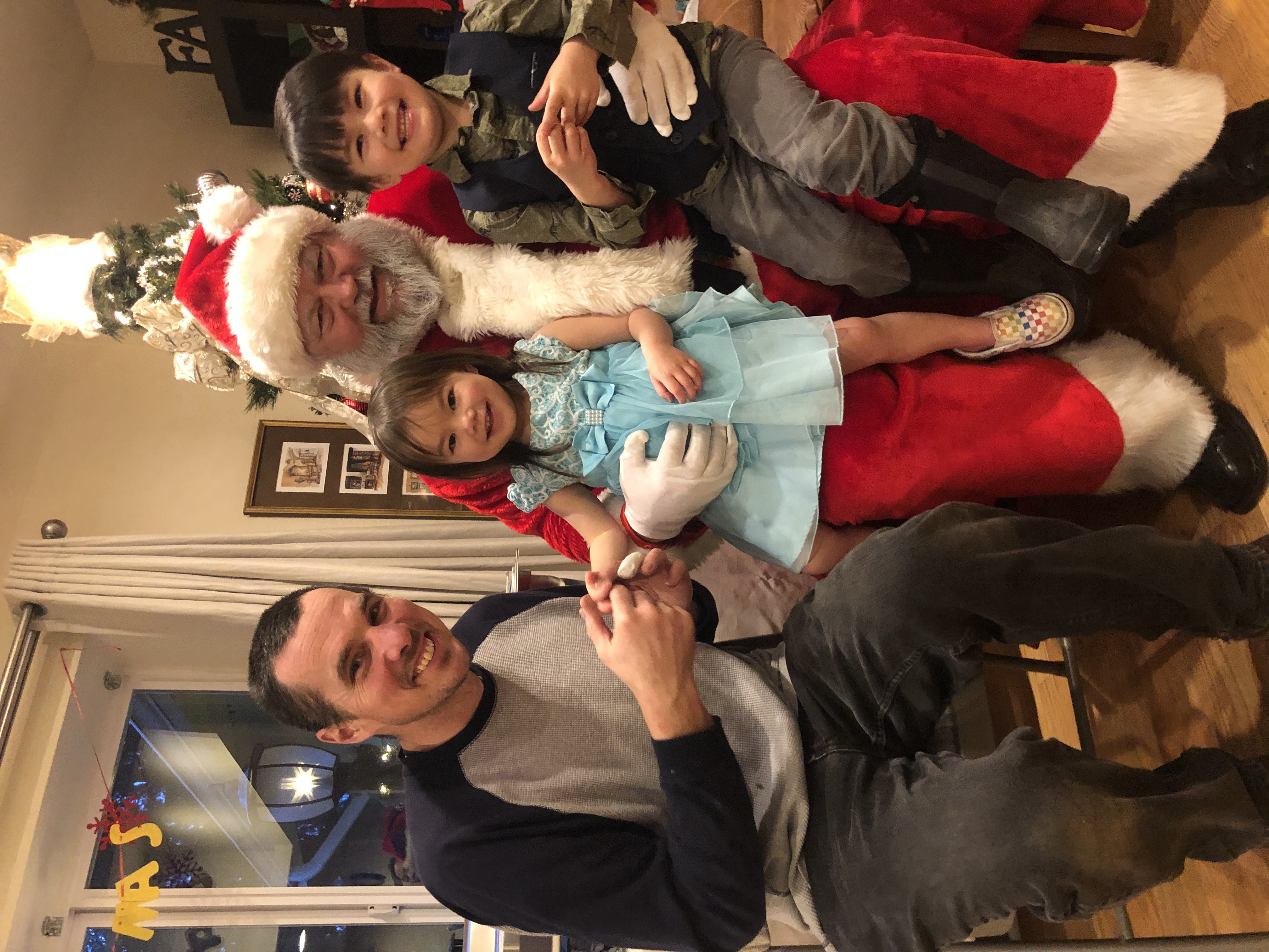 Man and two smiling children sitting with a person dressed as Santa Claus in a decorated room with a Christmas tree.