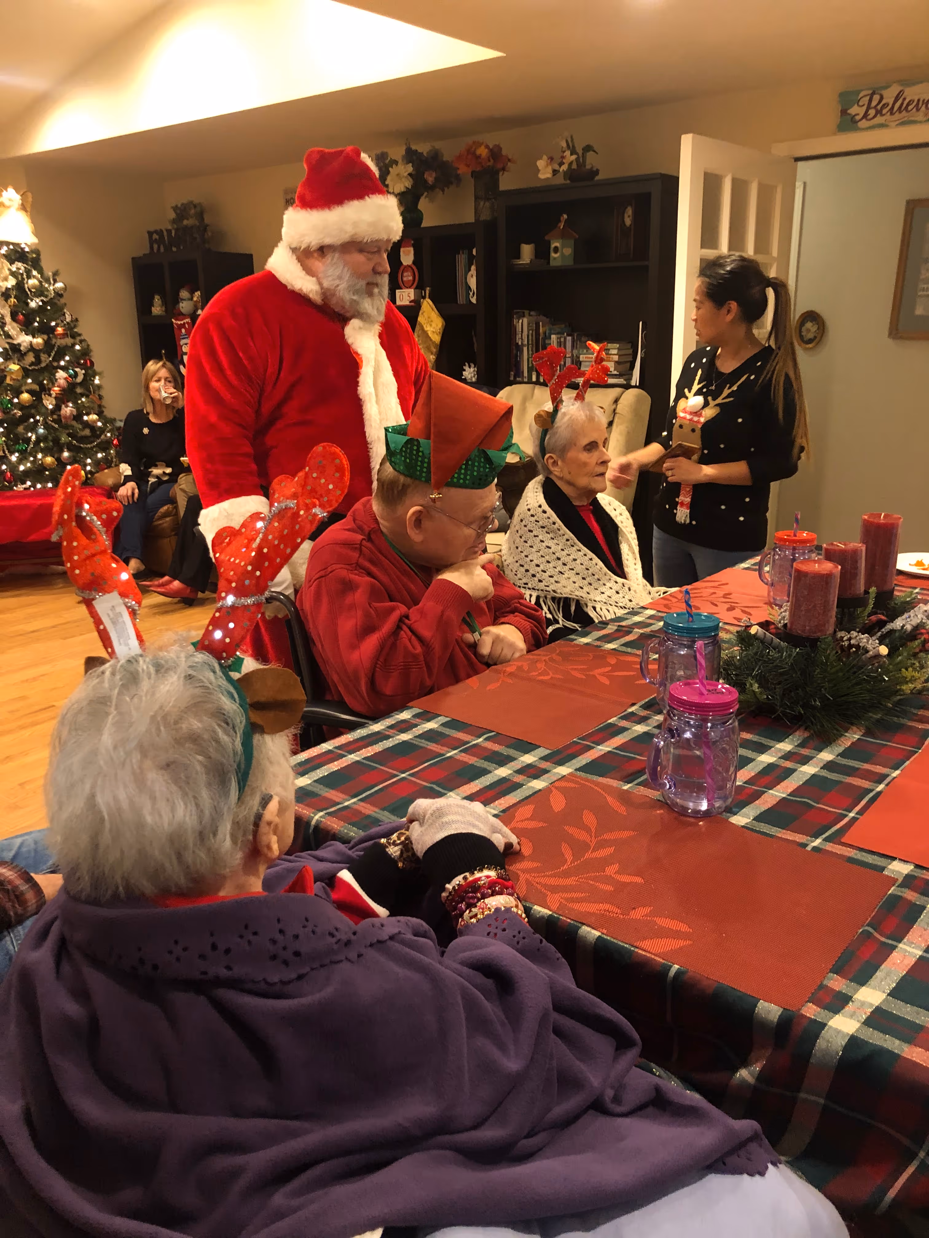 Group of elderly people wearing Christmas accessories sitting around a festively decorated table with a man dressed as Santa Claus standing nearby.