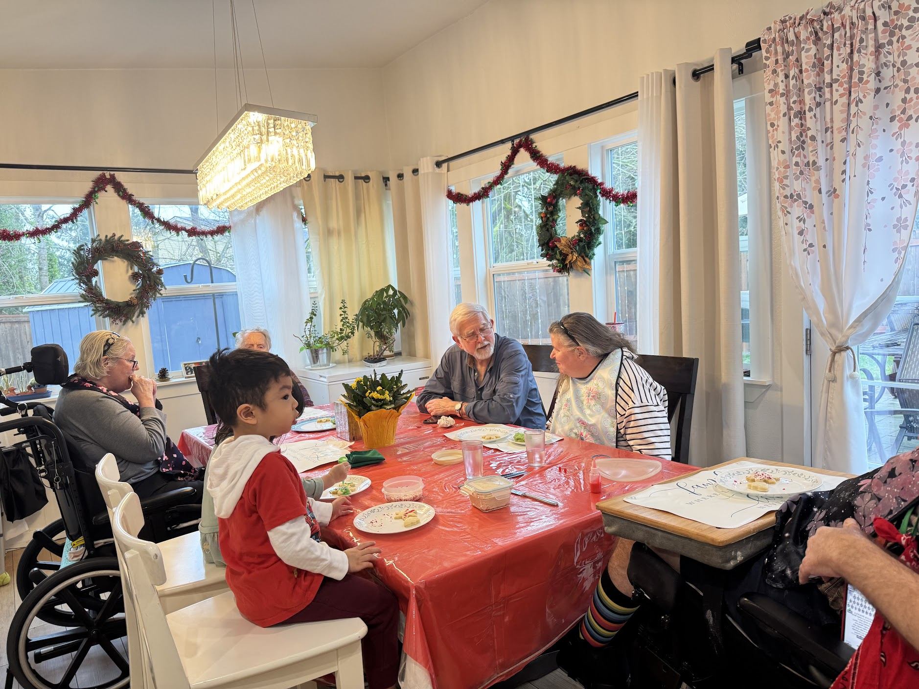 A multi-generational group seated around a dining table with red tablecloth, decorating and eating cookies in a cozy room with holiday wreaths and garlands on the windows.